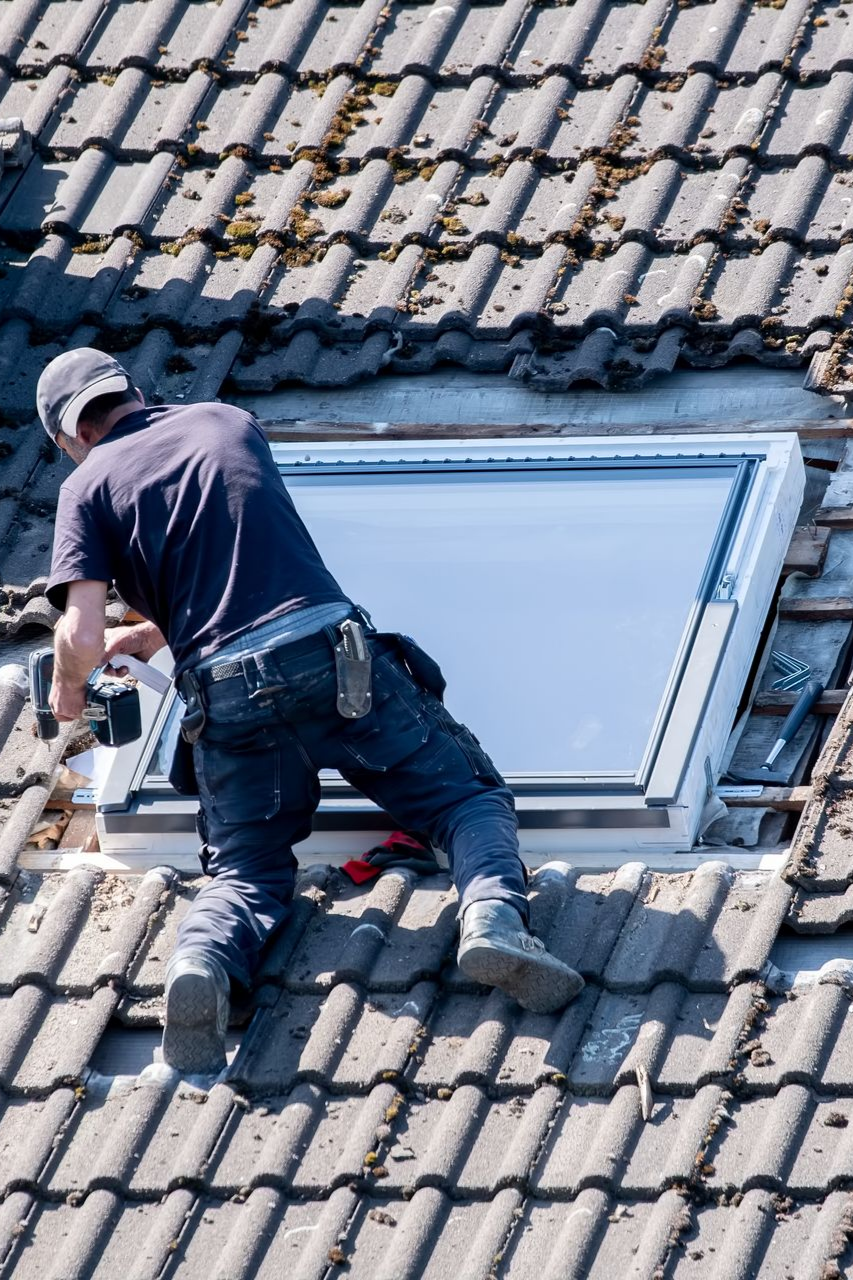 Un couvreur installe un puits de lumière sur un toit en tuile à l'aide d'une perceuse électrique.