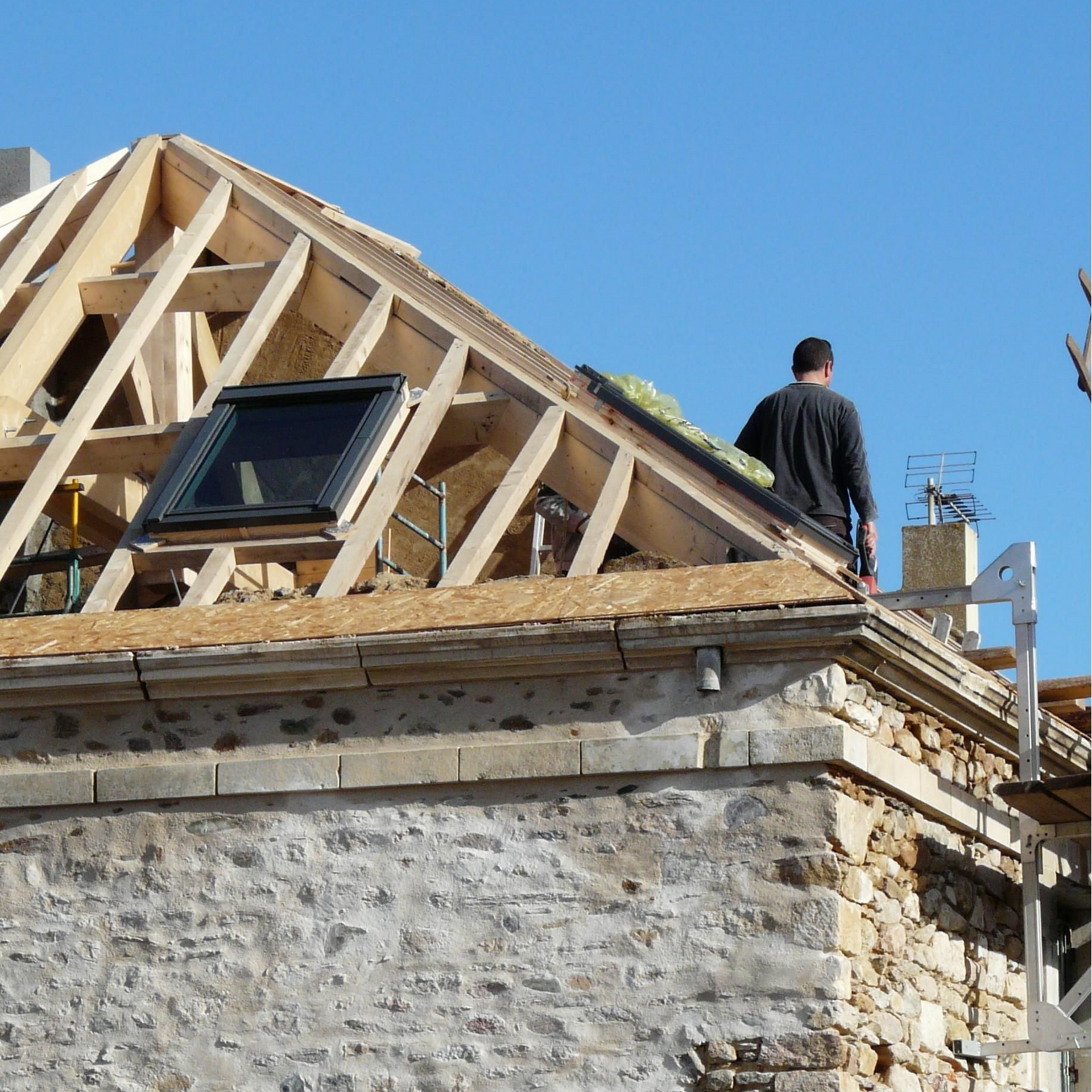 Rénovation de charpente : homme sur un toit avec poutres en bois apparentes et puits de lumière.