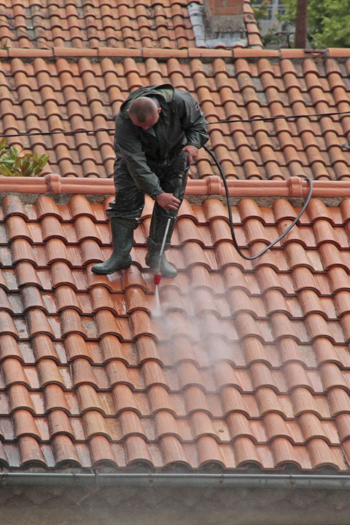 Une personne en combinaison verte nettoie au nettoyeur à haute pression un toit de tuile rouge.