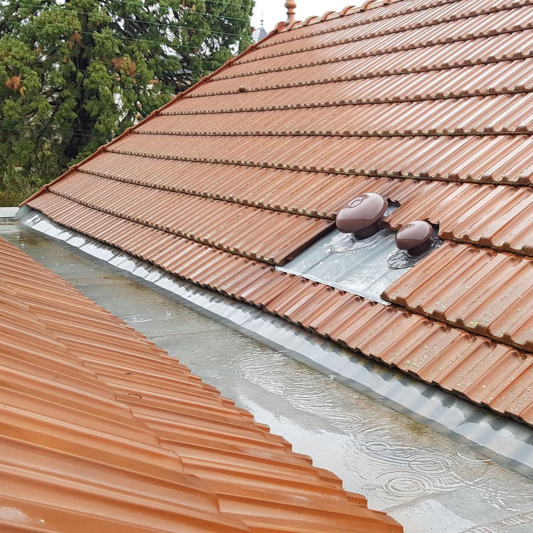 Toiture en tuiles avec deux aérations rondes, eau de pluie s'accumulant dans une gouttière métallique.