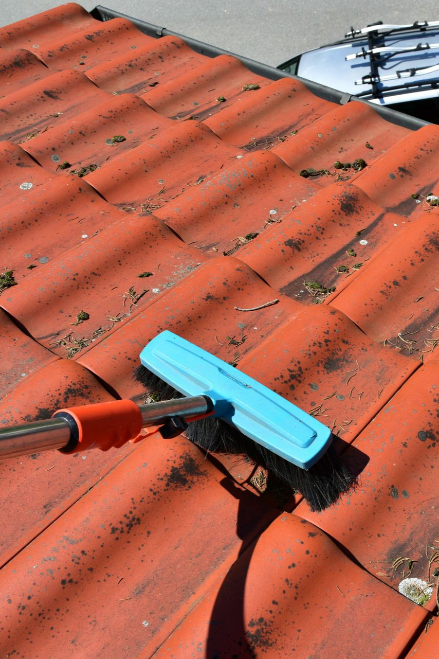 Une personne nettoie un toit de tuile rouge avec une brosse bleue à long manche.