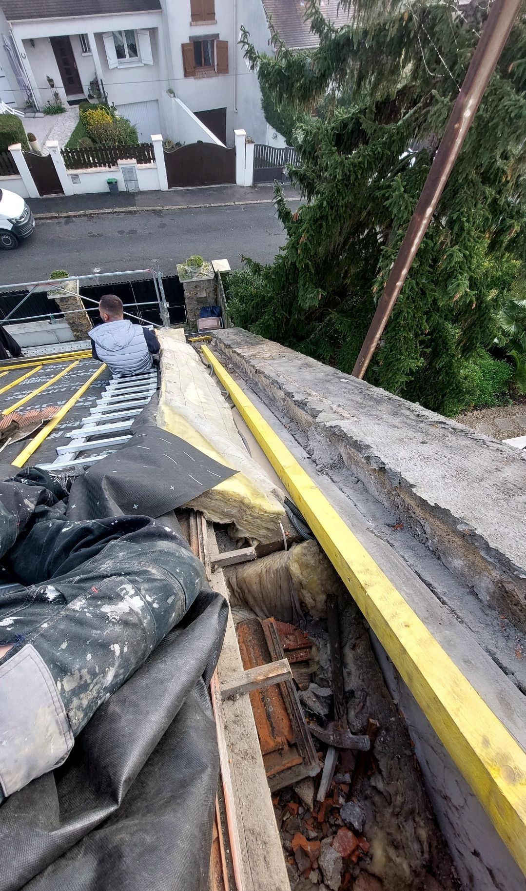 Travaux de toiture : Une personne travaille sur un toit endommagé à l’aide de matériaux et d’une poutre jaune. Des maisons et une voiture sont visibles en arrière-plan.