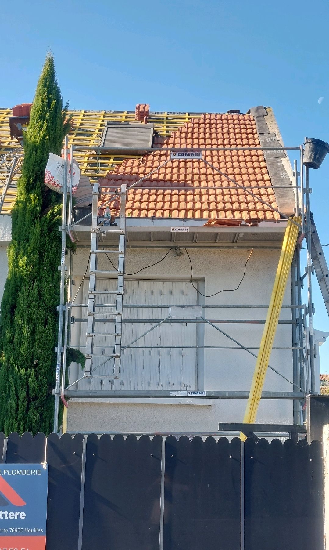 Maison en réparation de toiture avec échafaudages ; tuiles rouges, échelle jaune, ciel bleu.