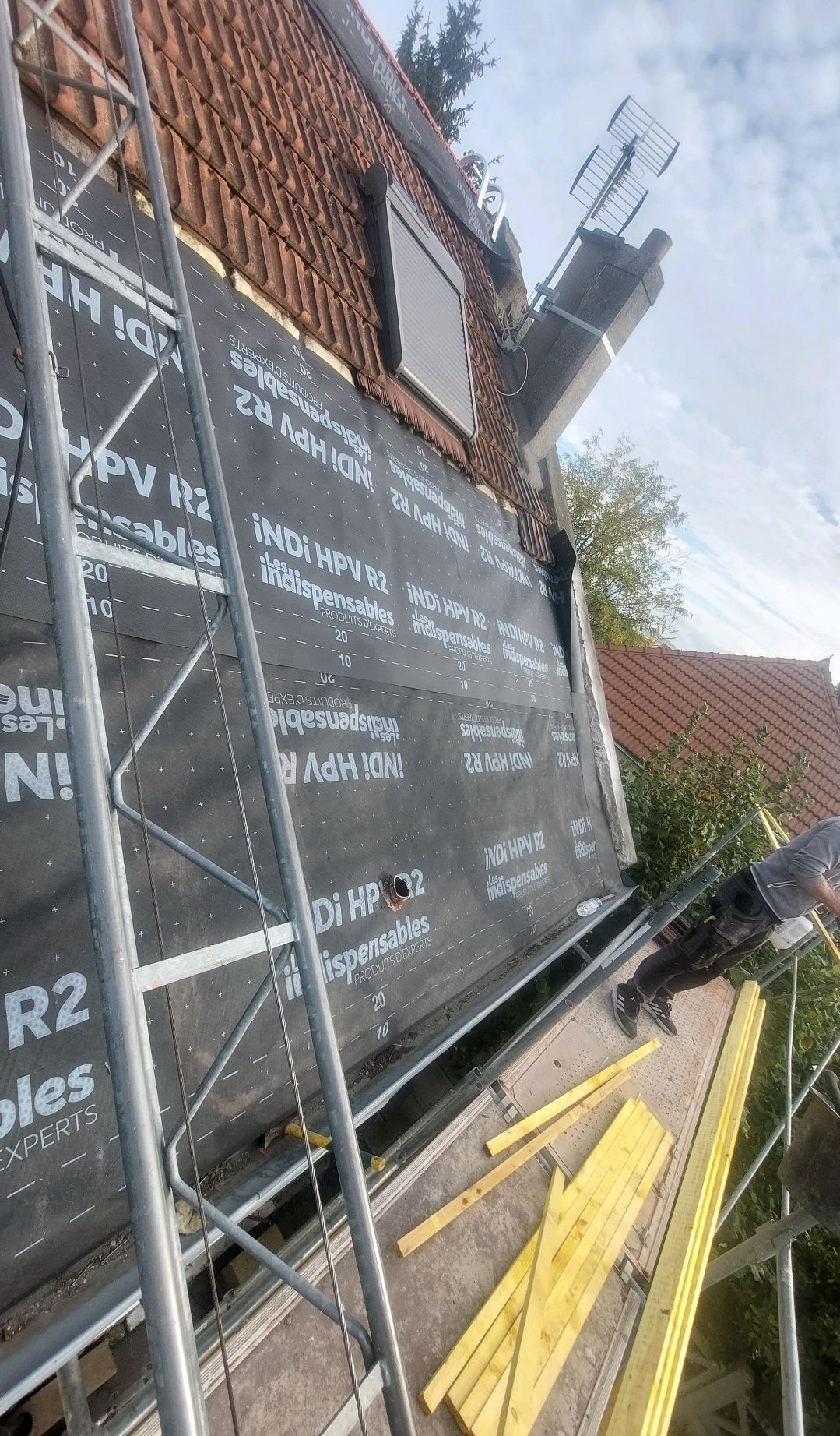 Couvreur sur échafaudage posant une membrane d'étanchéité noire sur un toit de tuiles rouges. Lumière du soleil, extérieur.