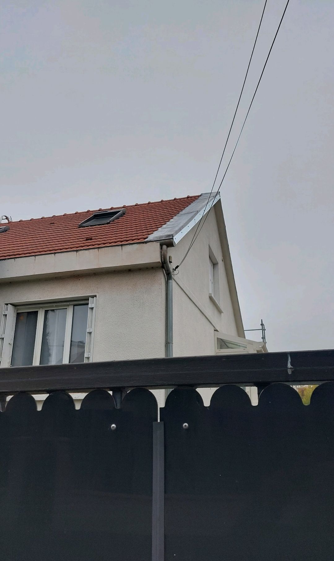 Maison au toit de tuiles rouges, clôture noire et lignes électriques se détachant sur un ciel nuageux.