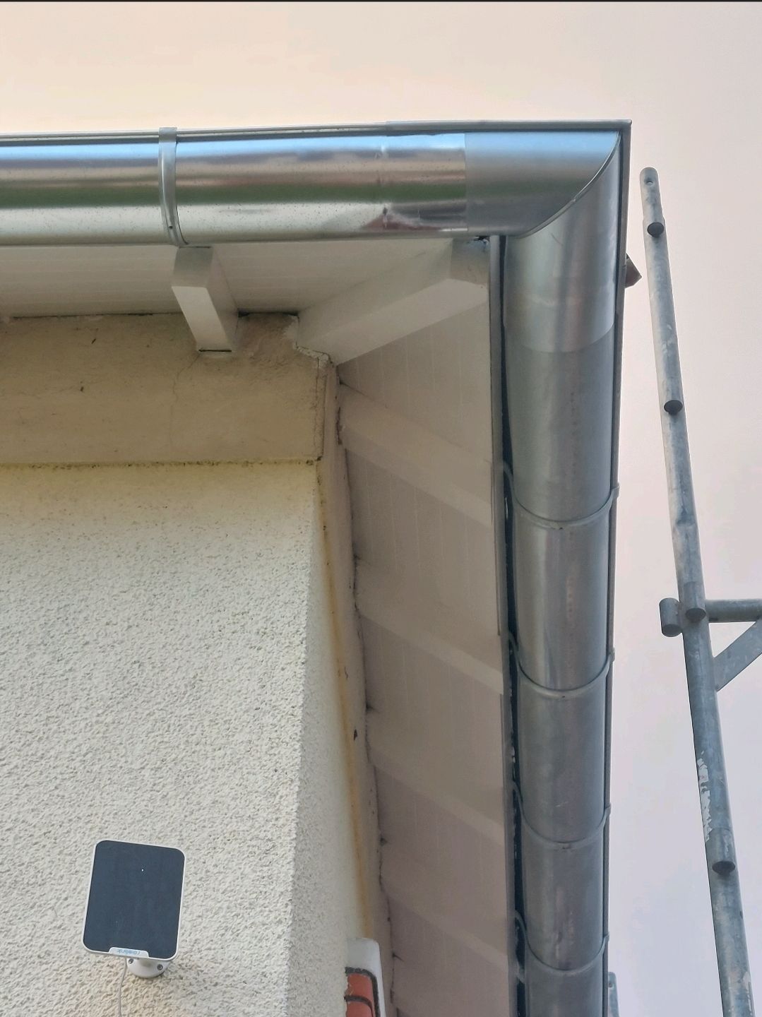 Coin d'un bâtiment aux gouttières argentées. Une échelle se trouve à côté de la gouttière.