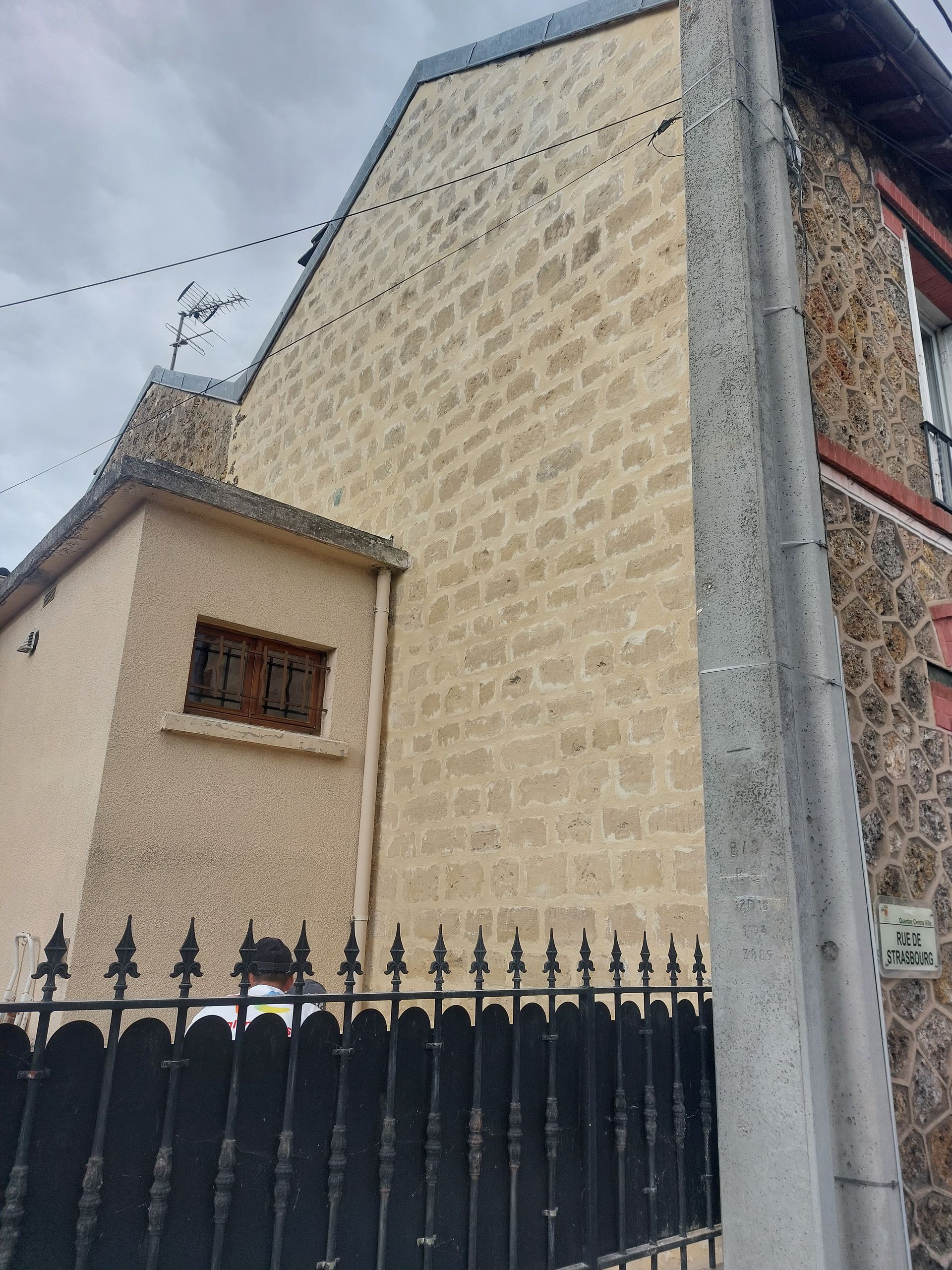 Extérieur du bâtiment avec murs en pierre et en stuc, clôture en fer forgé noir et ciel couvert.
