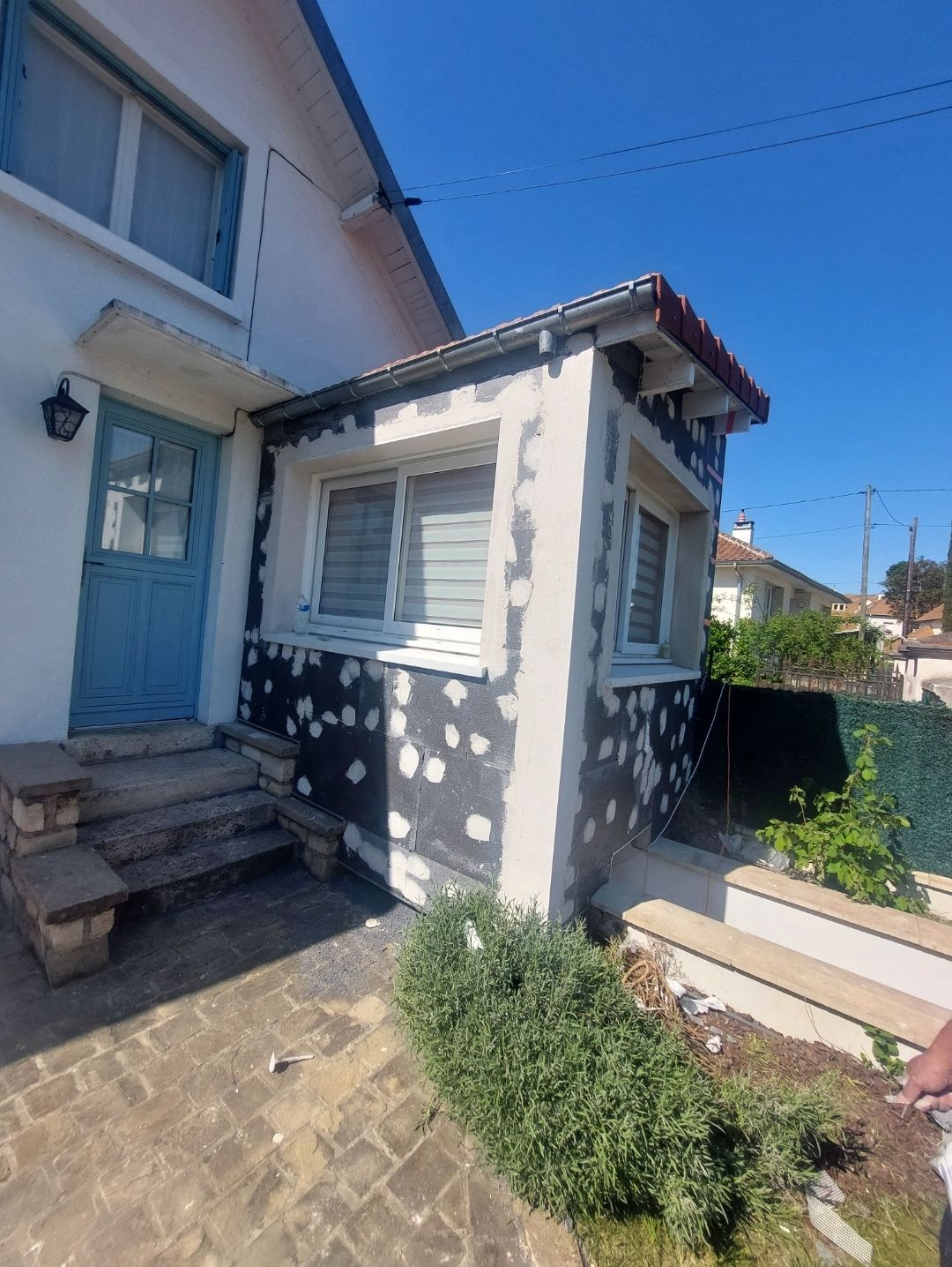 Extérieur d'une maison en construction par une journée ensoleillée, avec une porte bleue, des fenêtres blanches et un bardage gris.