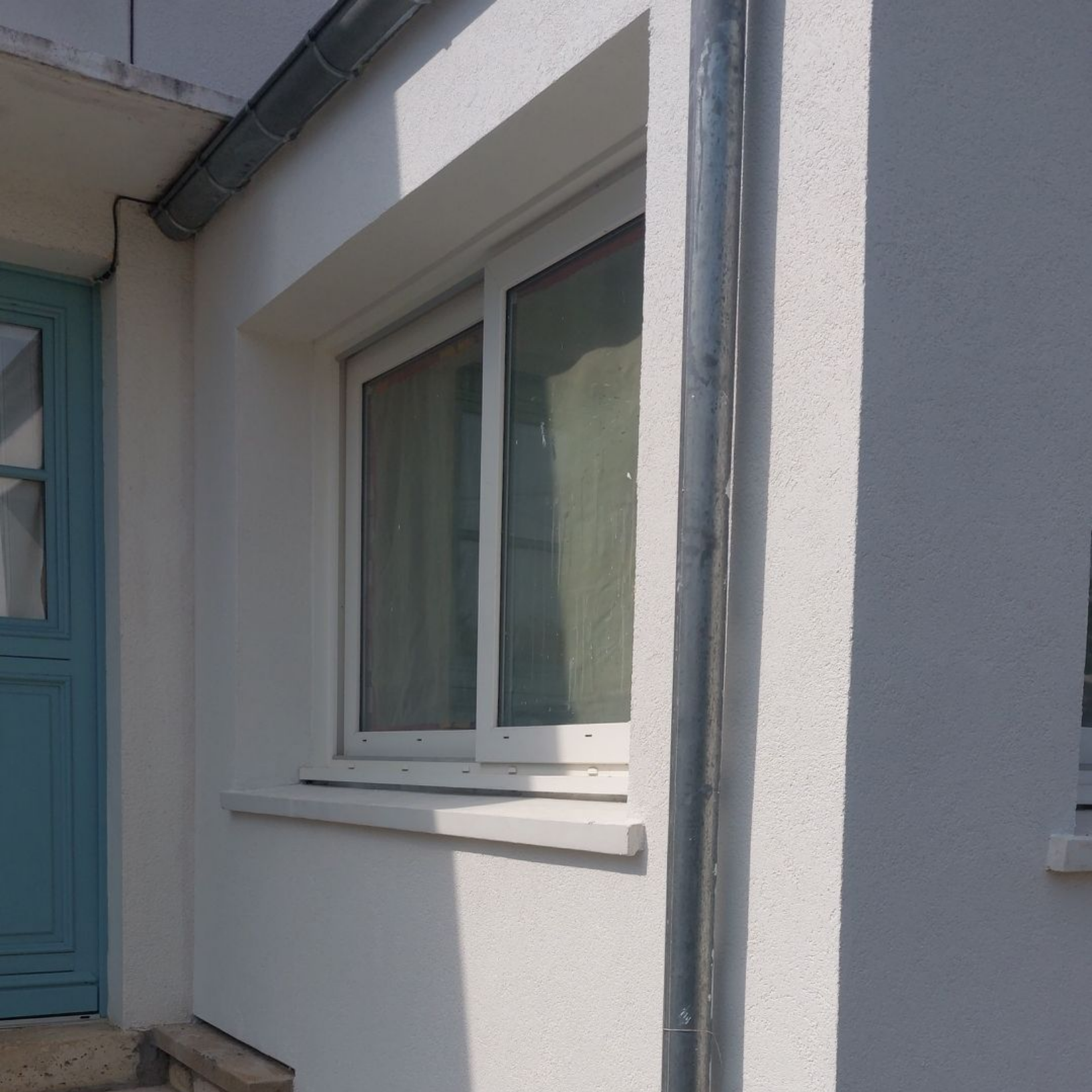 Fenêtre et moulures blanches sur un mur extérieur blanc, à côté d'une porte turquoise, avec une gouttière argentée.