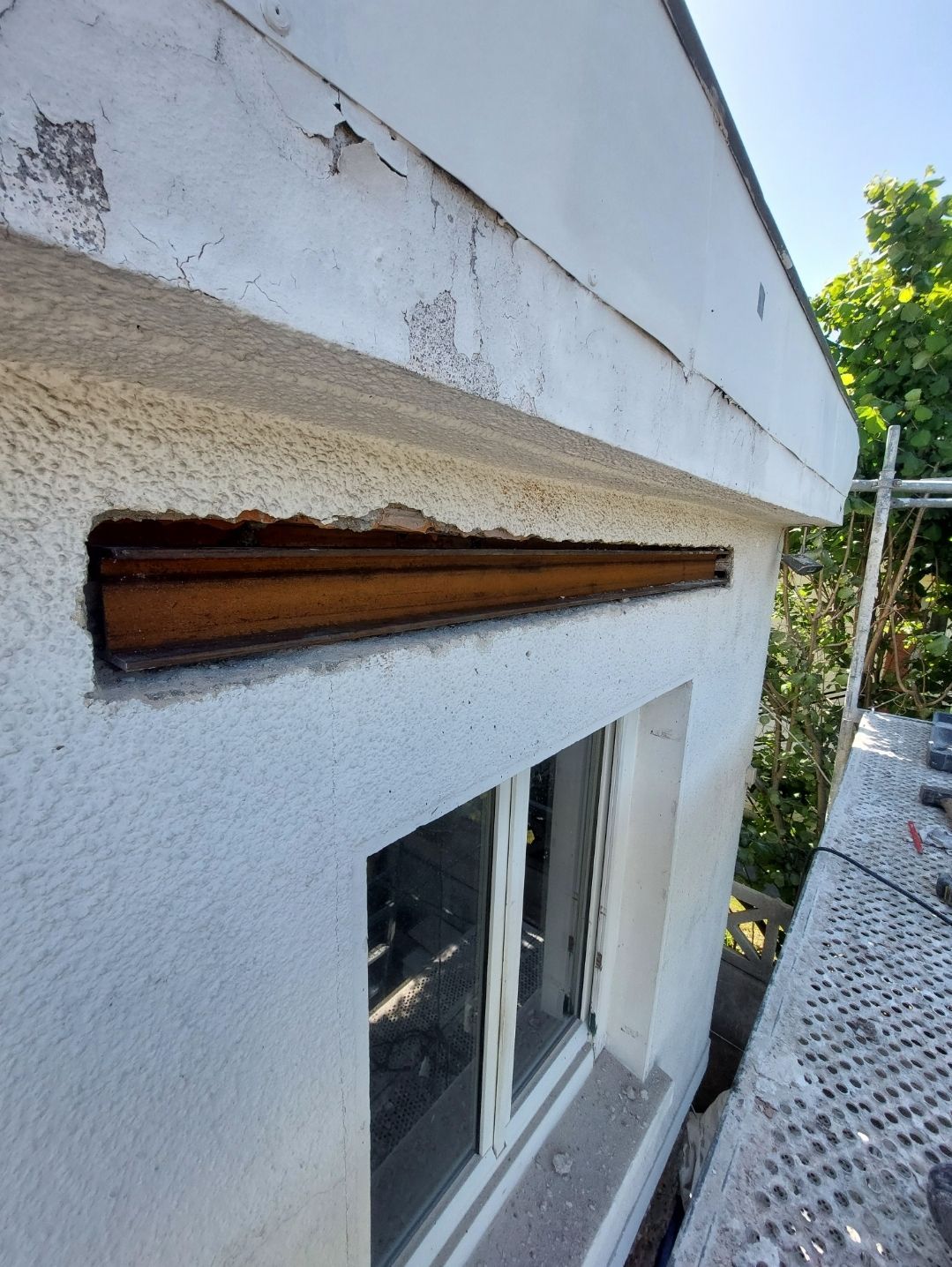 Vue extérieure d'un bâtiment avec une poutre en acier visible au-dessus d'une fenêtre, du béton et des murs blancs.