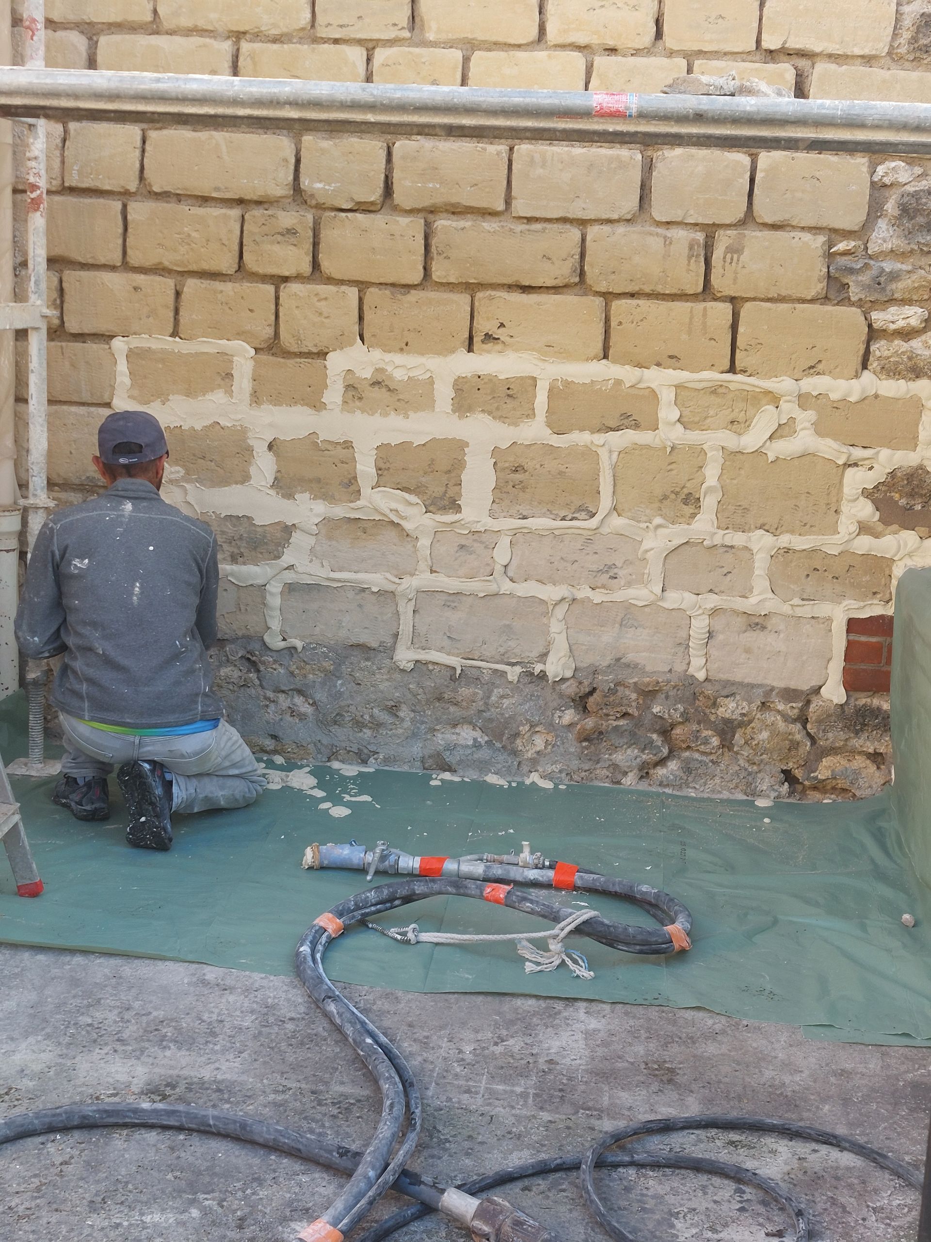 Une personne applique du plâtre sur un mur de briques. On aperçoit des échafaudages, des tuyaux d'arrosage et du matériel de plâtrage.