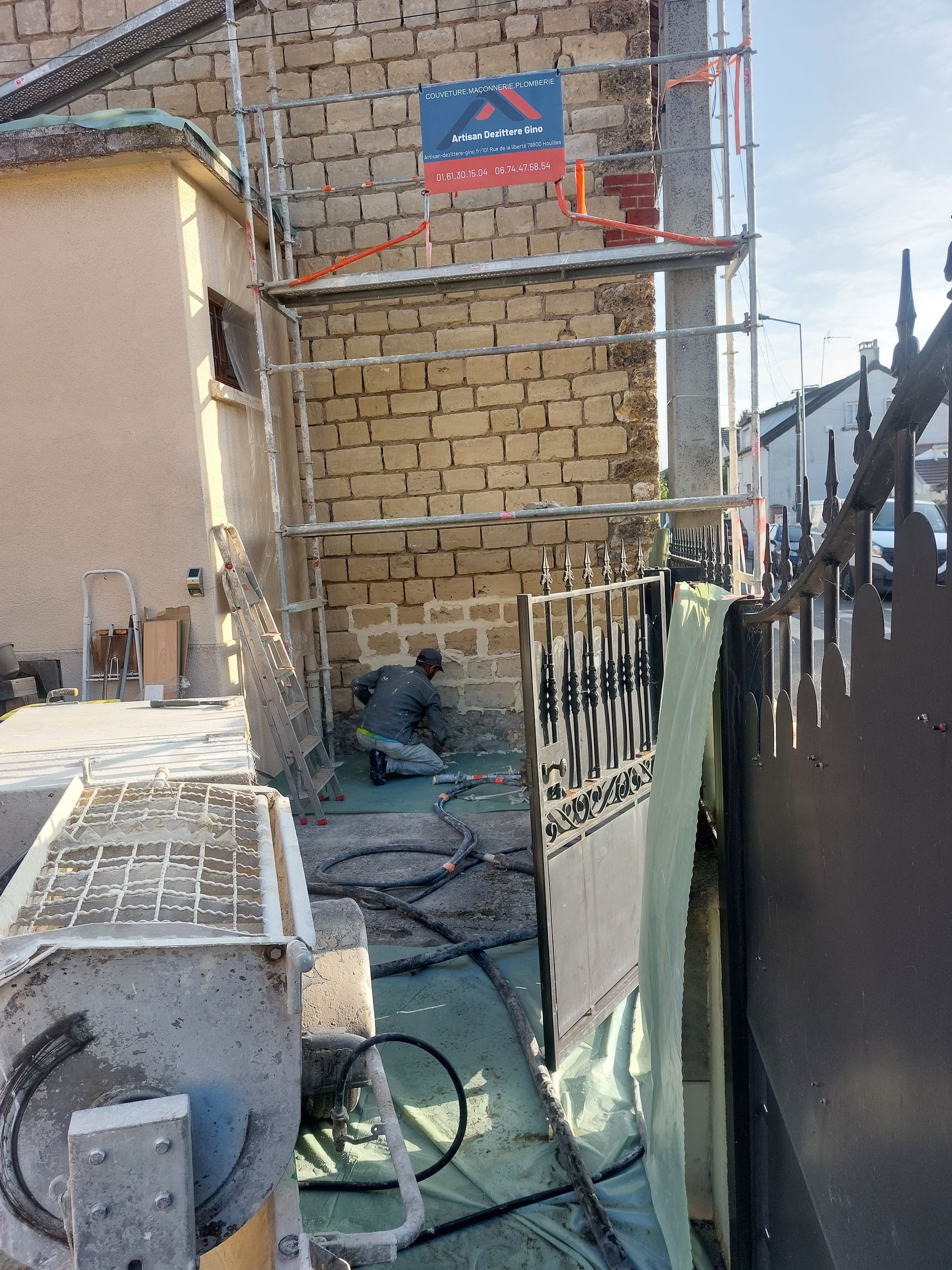 Un homme travaille sur la façade d'un bâtiment à l'aide d'échafaudages. Un portail noir se trouve au premier plan. Un mur de briques complète le tableau.