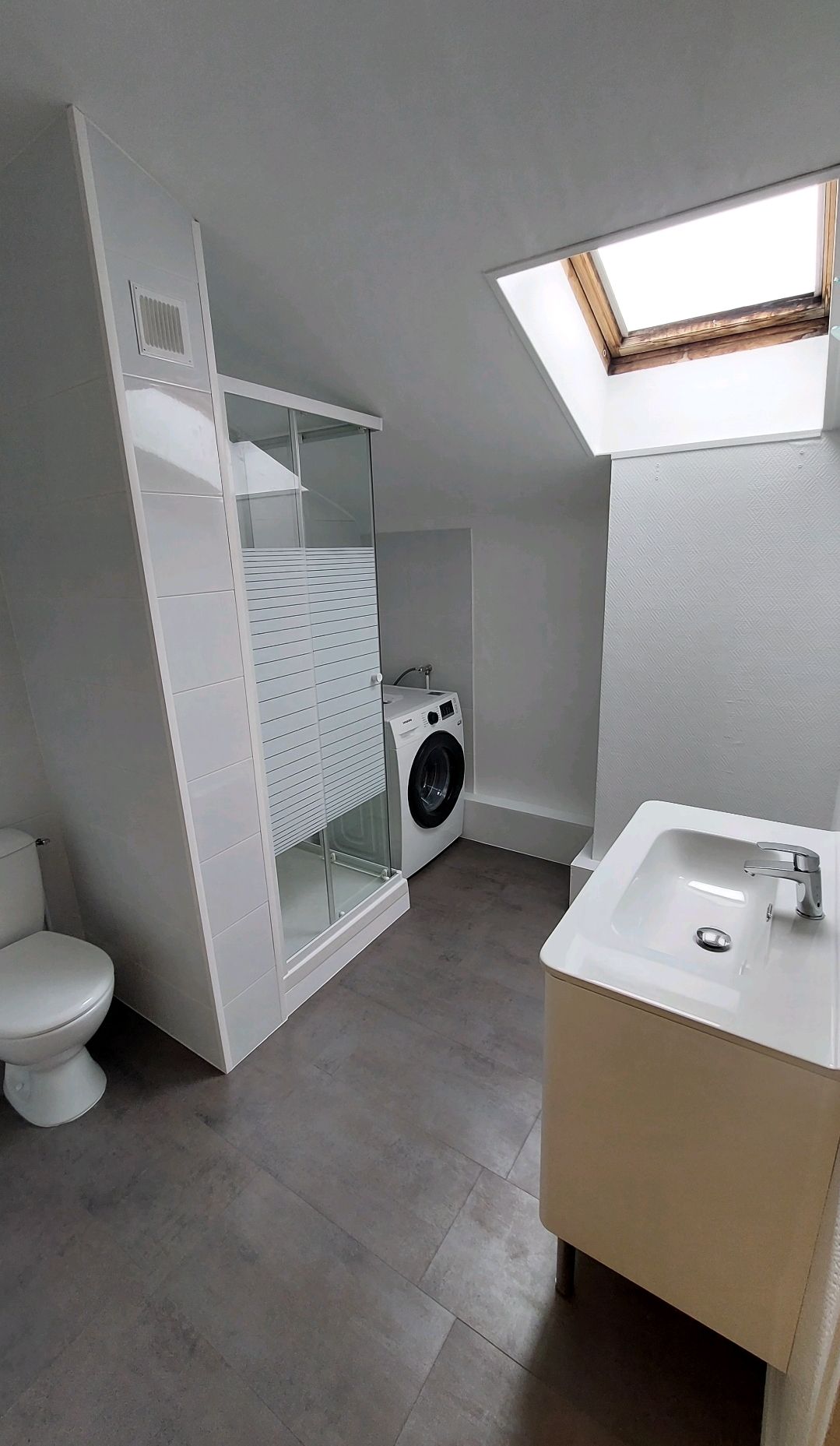Salle de bains avec toilettes, lavabo, lave-linge et puits de lumière. Sol gris, murs et placards blancs.