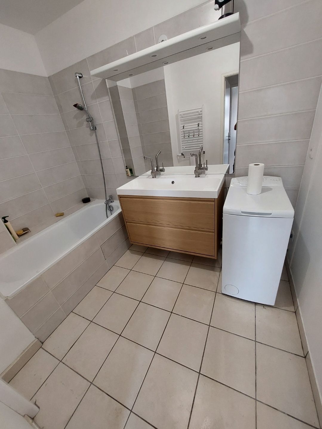 Salle de bains avec baignoire, double vasque et lave-linge. Sols et murs carrelés clairs.