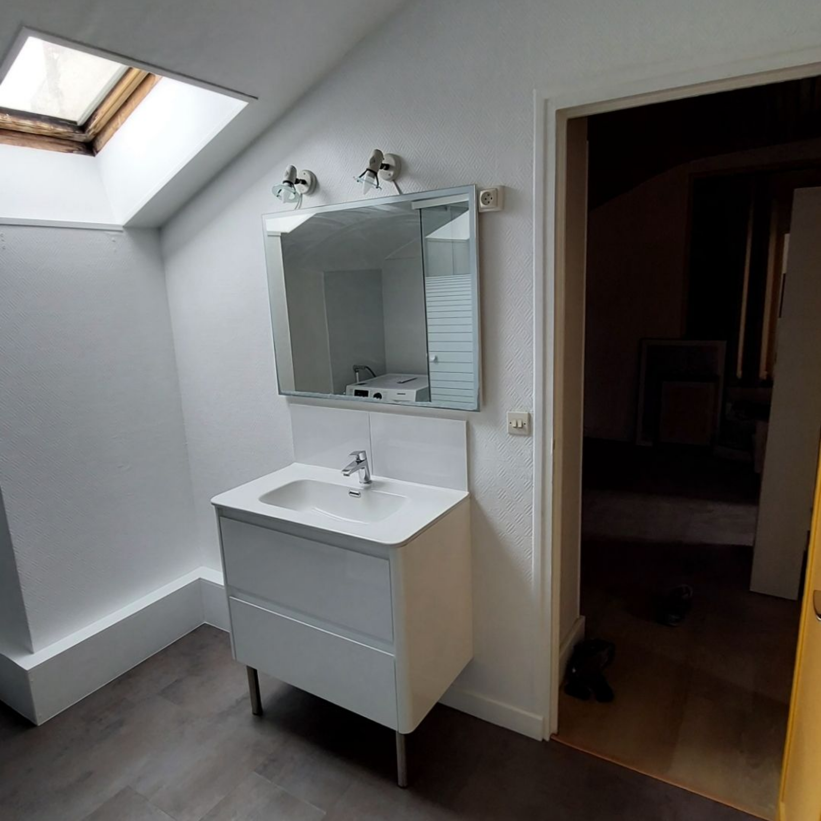 Salle de bains blanche avec lavabo, miroir et puits de lumière. Une porte ouverte est visible.