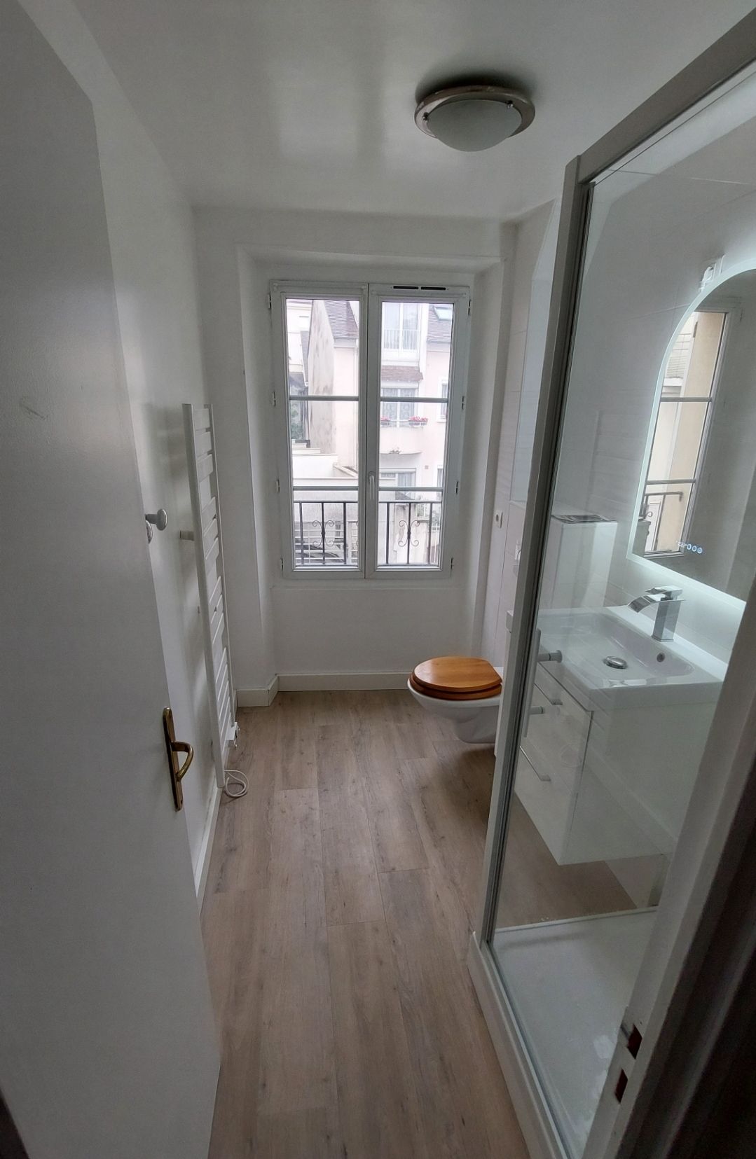 Salle de bains étroite avec fenêtre, toilettes, douche, lavabo et porte-serviettes.