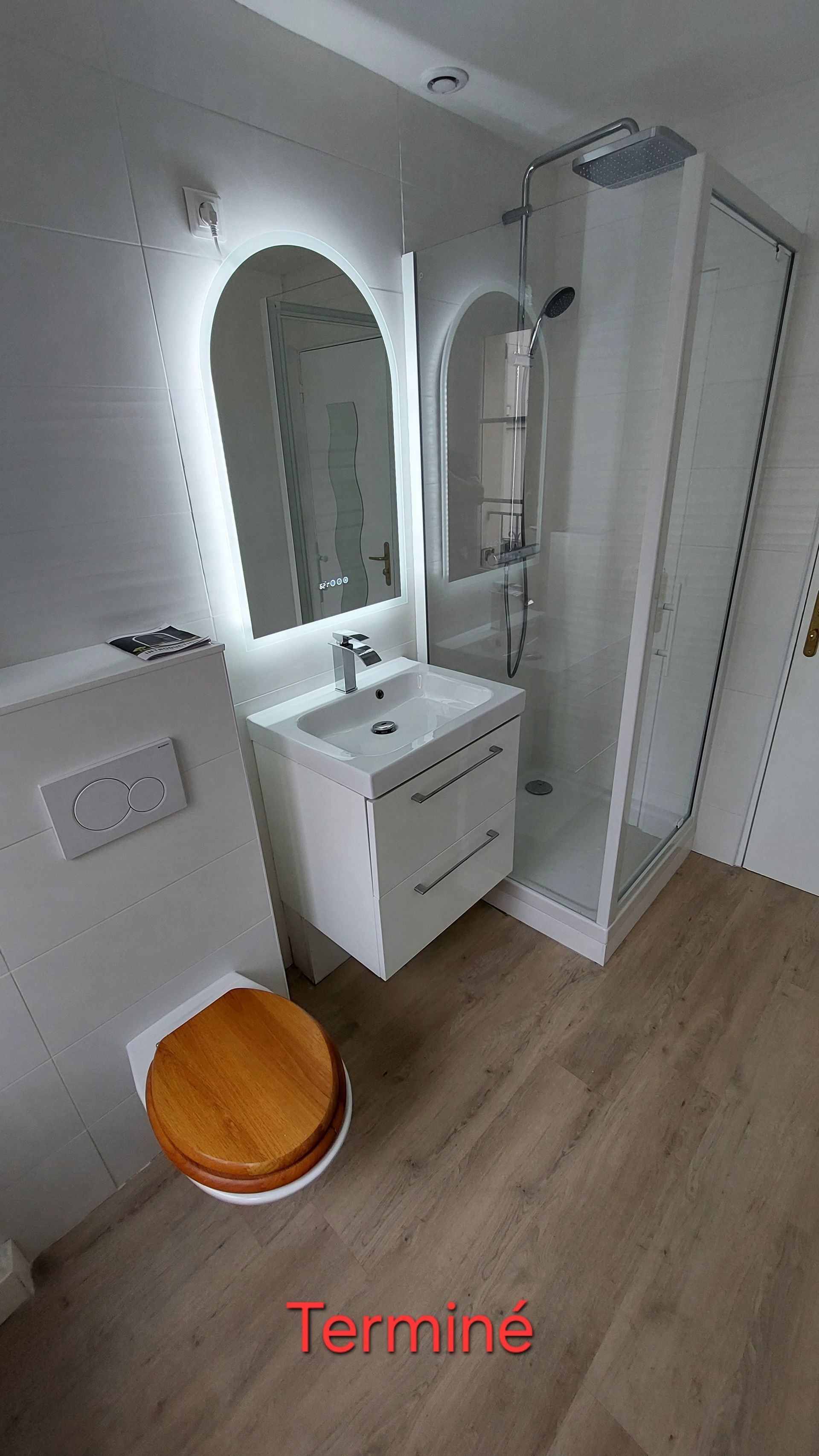 Petite salle de bains blanche avec douche, toilettes, lavabo et miroir. Sol clair imitation bois.