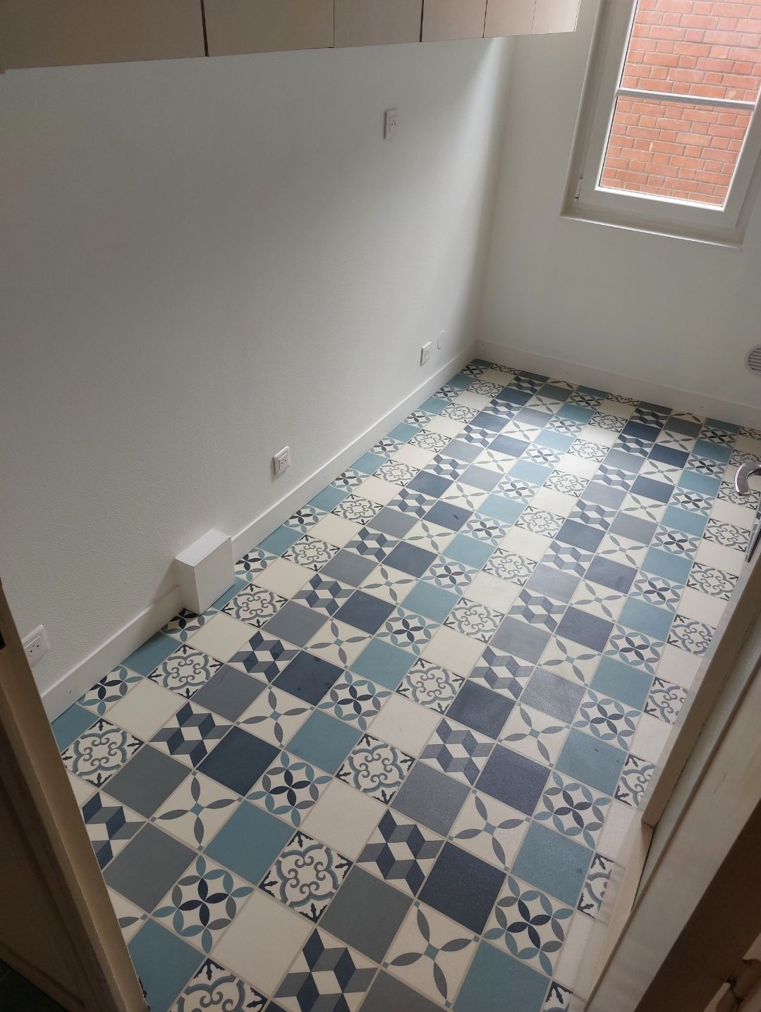 Chambre avec sol carrelé de carreaux bleus et blancs à motifs, murs blancs et petite fenêtre.