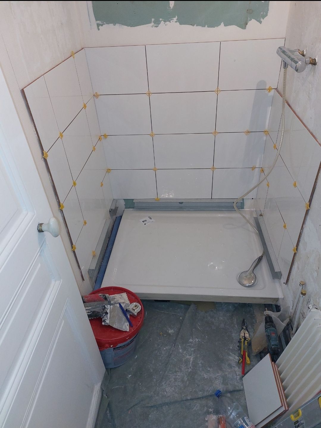 Salle de bains en construction : murs de douche carrelés en blanc, lavabo de douche, outils de carrelage et porte.
