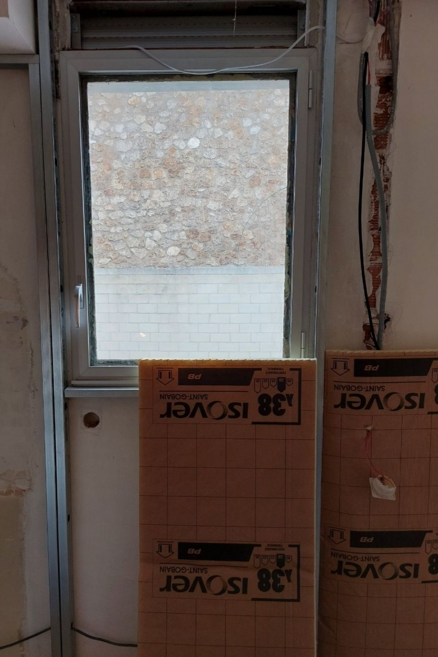 Fenêtre à cadre gris, entrouverte, laissant apparaître un mur de pierre à l'extérieur. Deux panneaux isolants rectangulaires sont placés sous la fenêtre.