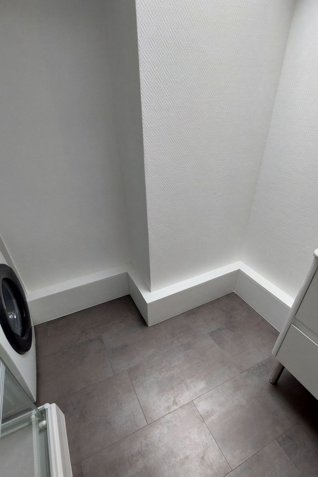 Coin buanderie avec machine à laver, sol carrelé foncé, murs et moulures blancs.