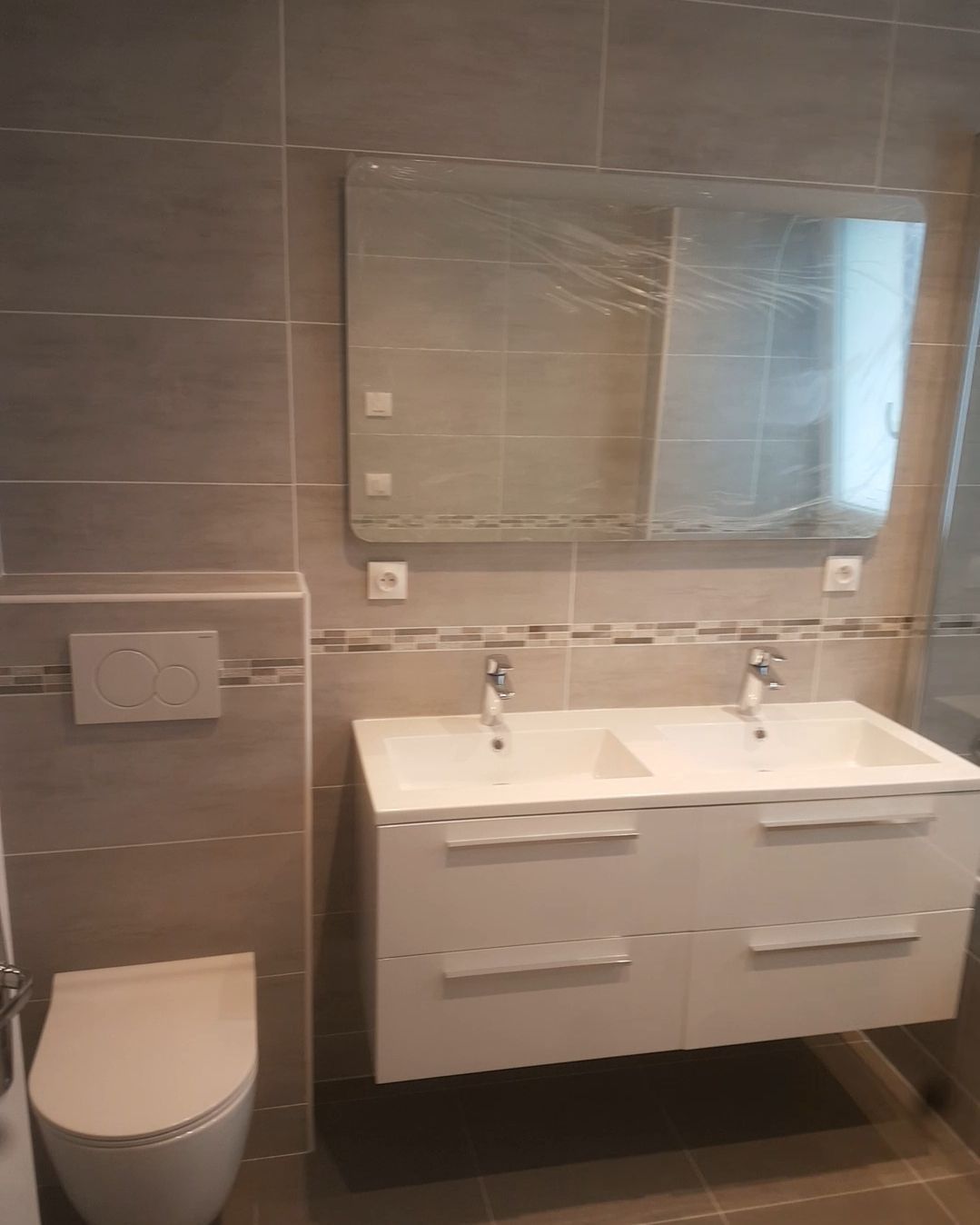 Salle de bains moderne avec double vasque, toilettes et grand miroir. Murs carrelés aux tons neutres.