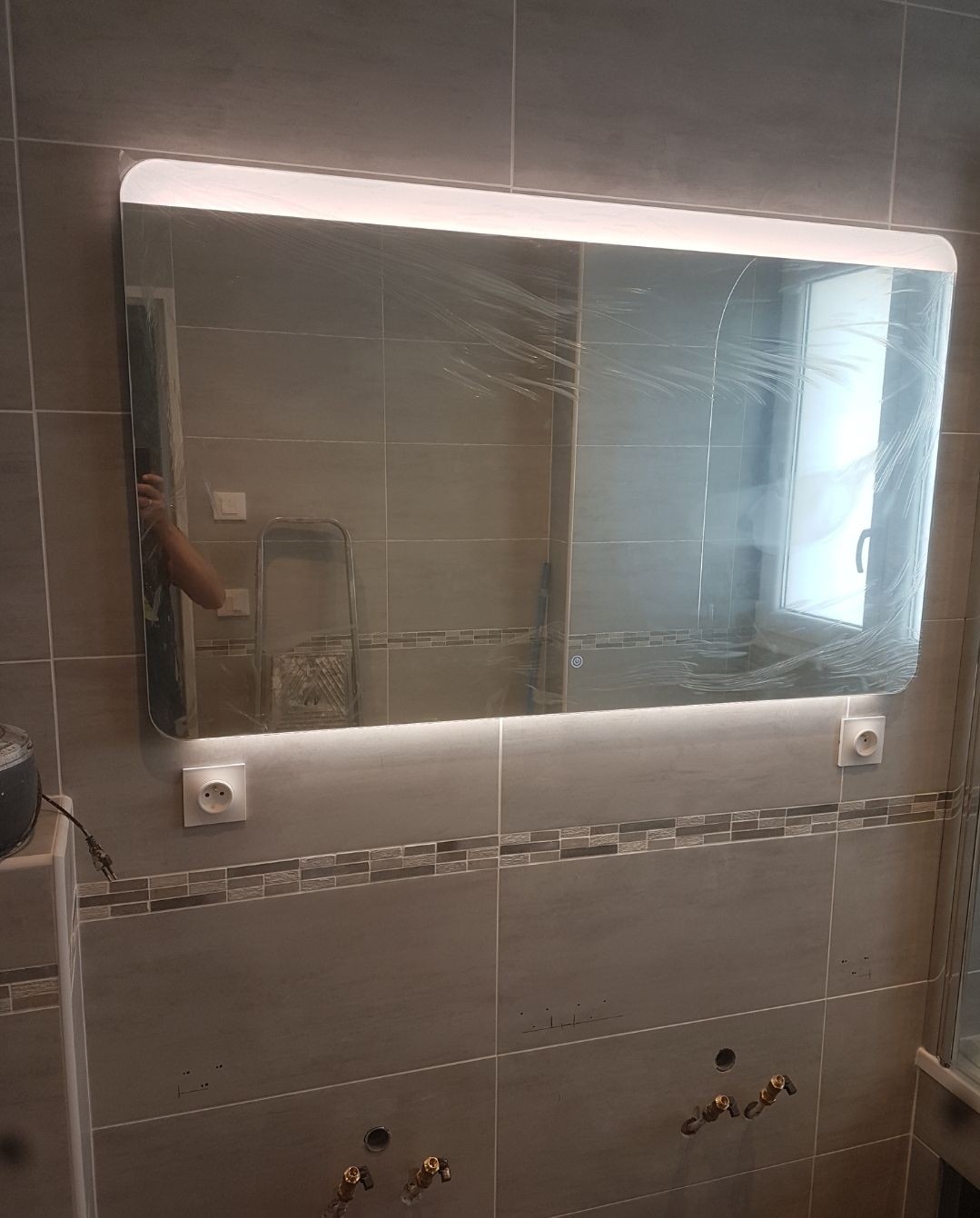 Miroir de salle de bains avec éclairage à LED intégré, reflétant la pièce, fixé sur un mur carrelé.