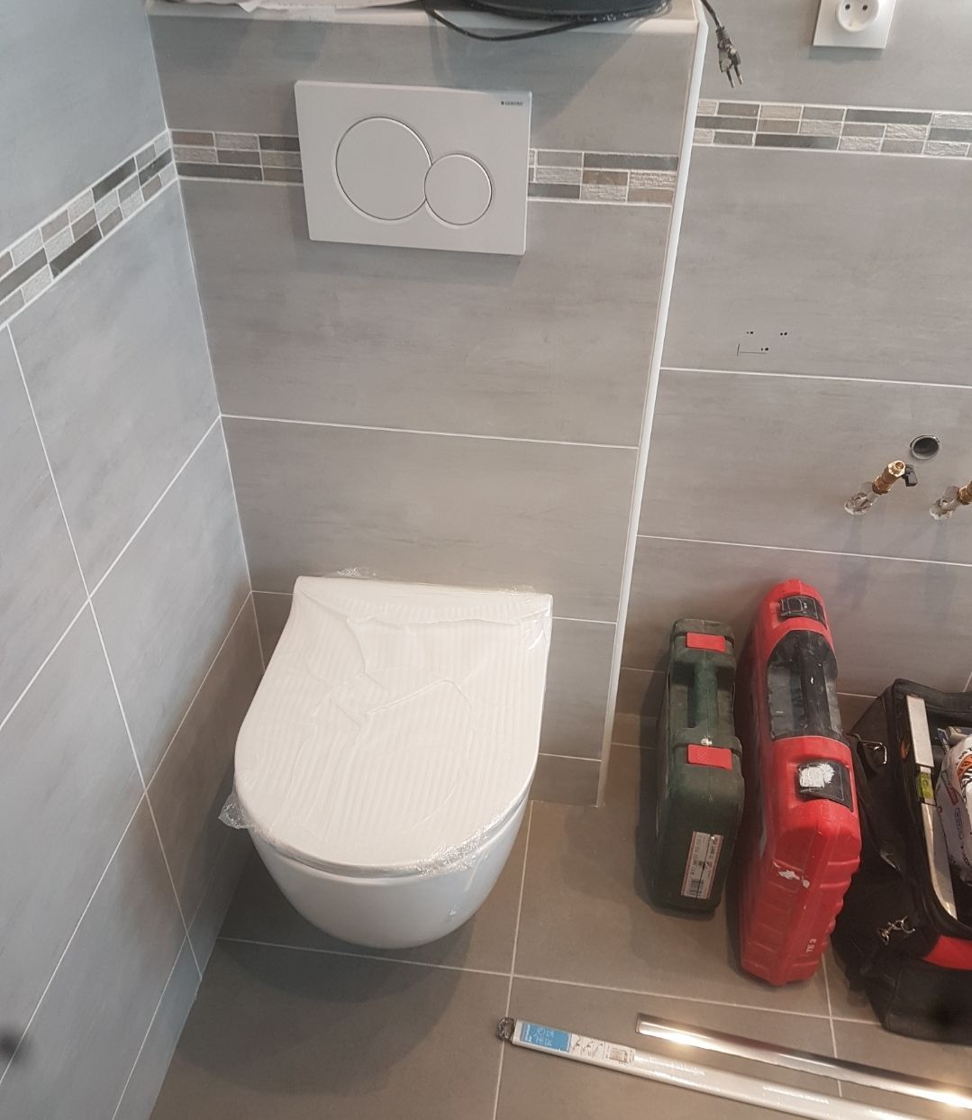 Des toilettes partiellement installées dans une salle de bains carrelée. Murs gris, toilettes blanches et accessoires au sol.