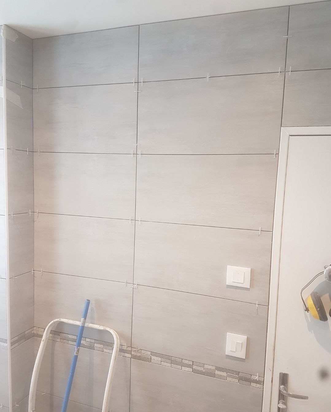 Mur carrelé gris avec des joints gris. Porte et interrupteurs blancs. Un escabeau se trouve dans un coin.