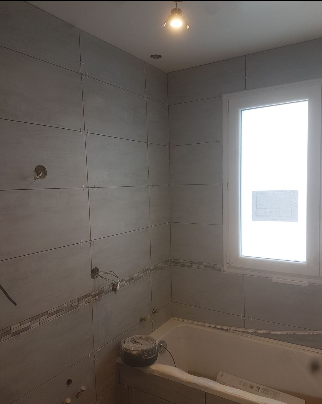 Rénovation de salle de bains en cours ; murs gris, fenêtre, baignoire. Câbles et installations électriques apparents.