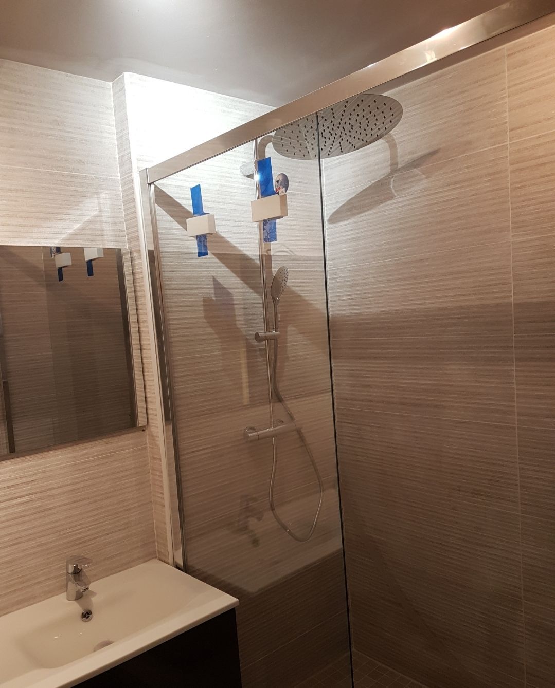 Salle de bains avec douche, porte en verre, pommeau de douche effet pluie, lavabo blanc et murs carrelés.