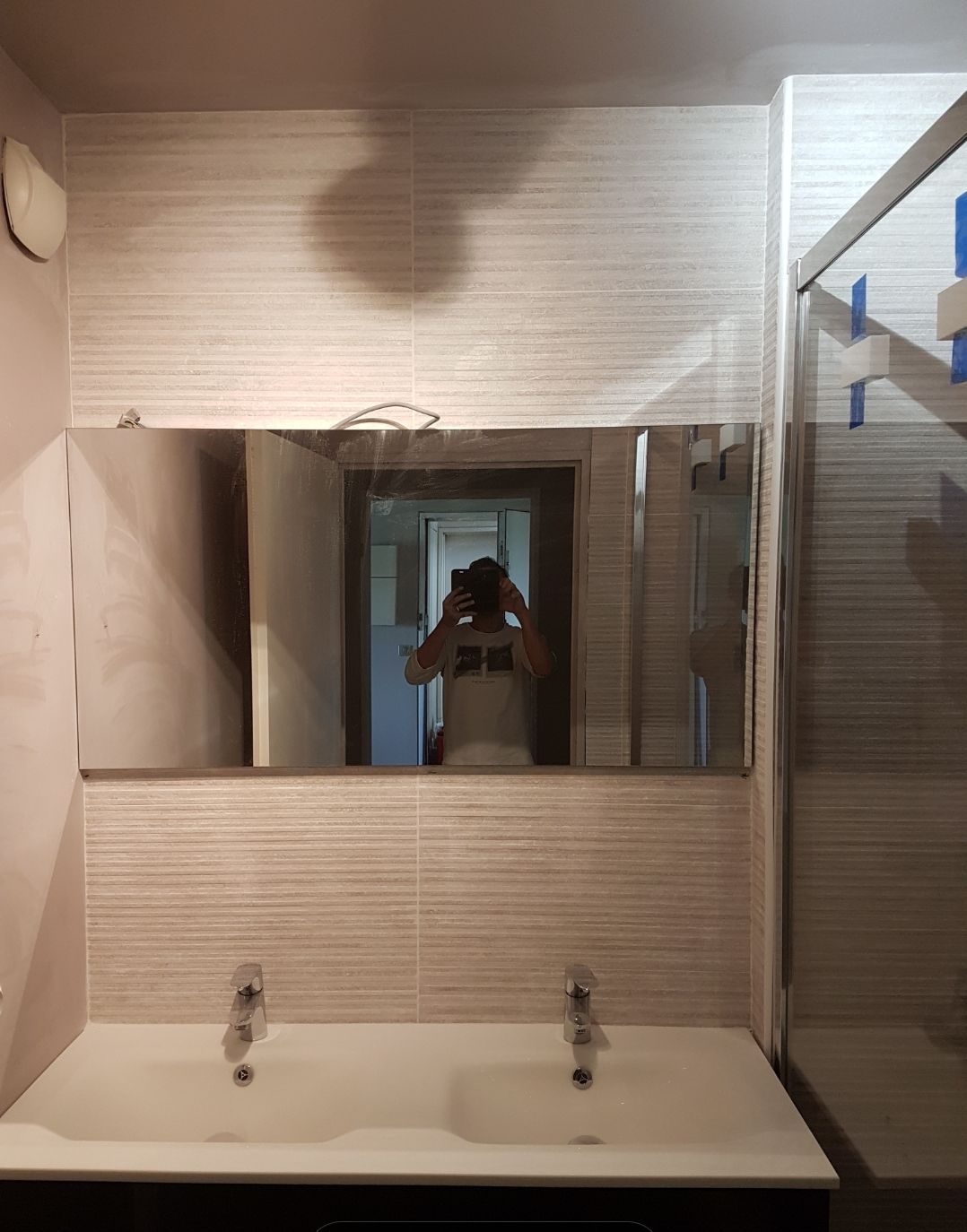 Salle de bains avec un miroir rectangulaire au-dessus d'un double lavabo, reflétant la personne qui prend la photo.