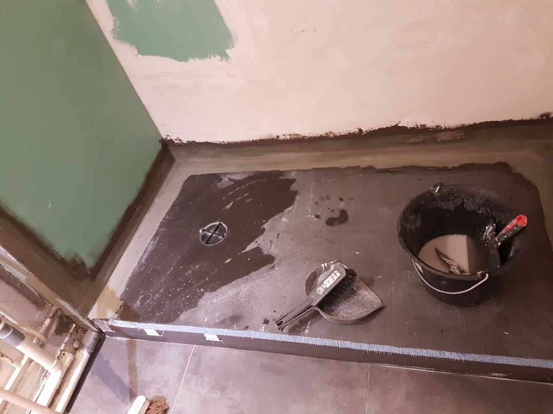 Receveur de douche en construction ; mastic gris foncé, seau noir avec outils, mur gris verdâtre, murs de couleur claire.