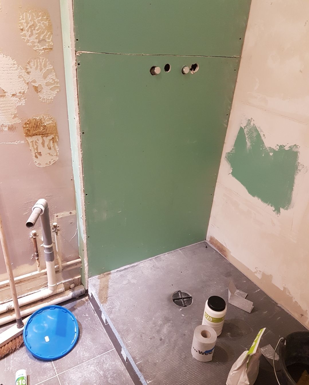 Rénovation de salle de bains en cours. Cloisons sèches vertes installées autour de la plomberie. Carrelage au sol, seaux et outils sur place.