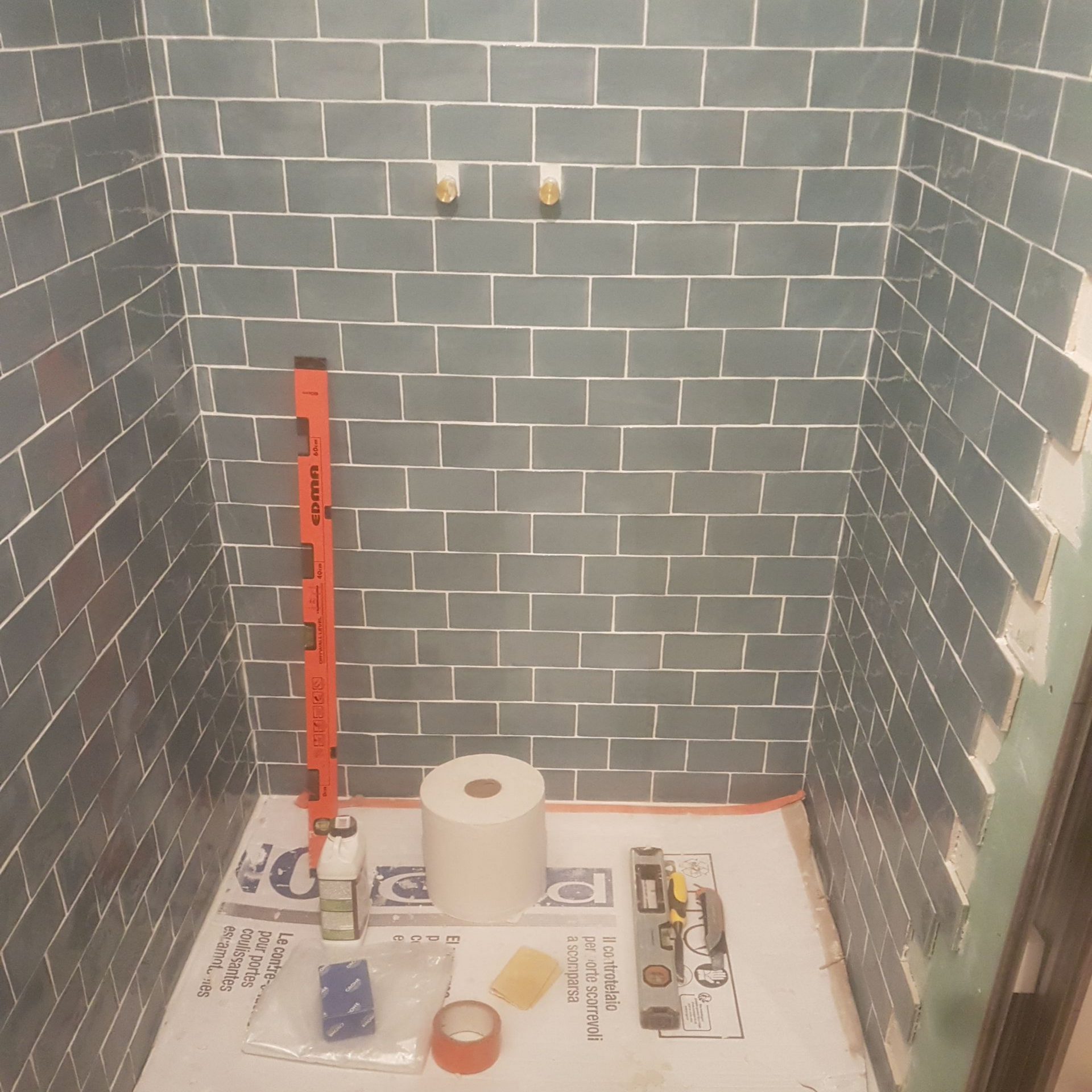 Une cabine de douche avec des carreaux et un sol blanc, un niveau orange, des produits de toilette et du papier toilette.