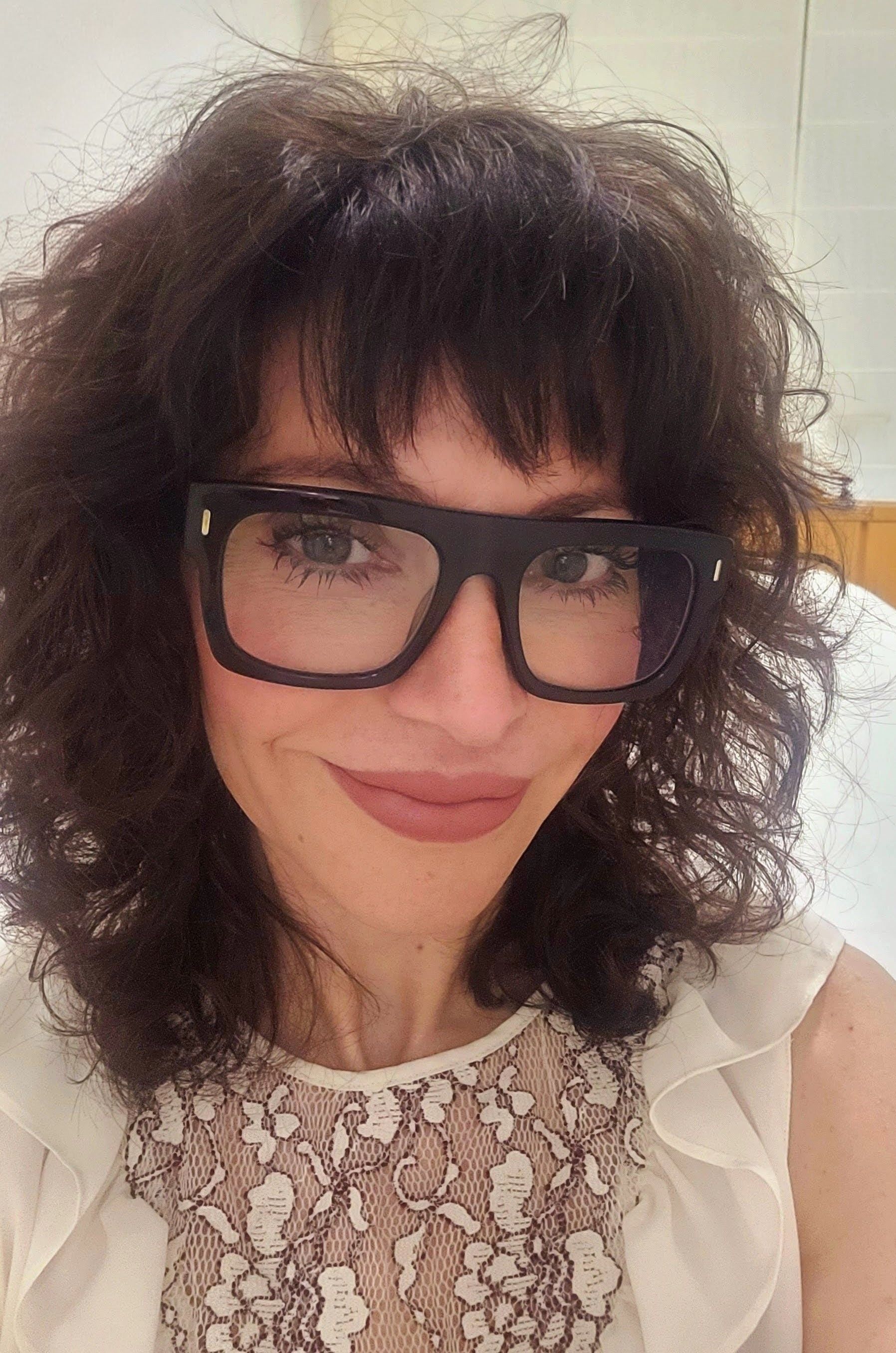 Mujer con cabello oscuro y rizado, con gafas, vestida con un top de color claro, mirando a la cámara.