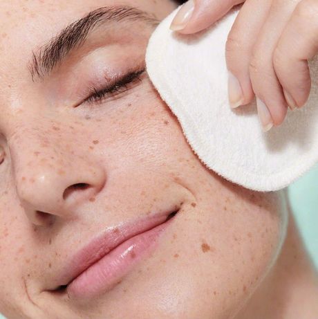 Mujer limpiando suavemente la cara con un disco de algodón blanco; primer plano, ojos cerrados, sonriendo.