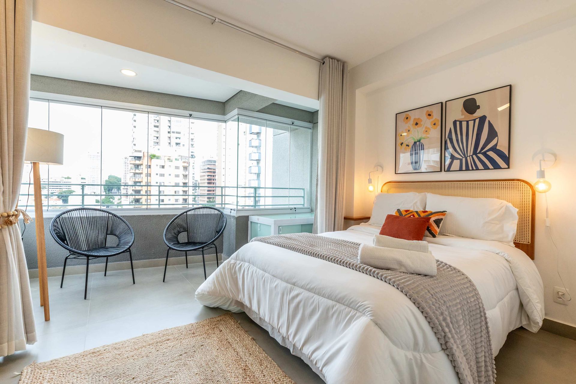 Bedroom with balcony: bed with white bedding, two chairs, and city view.