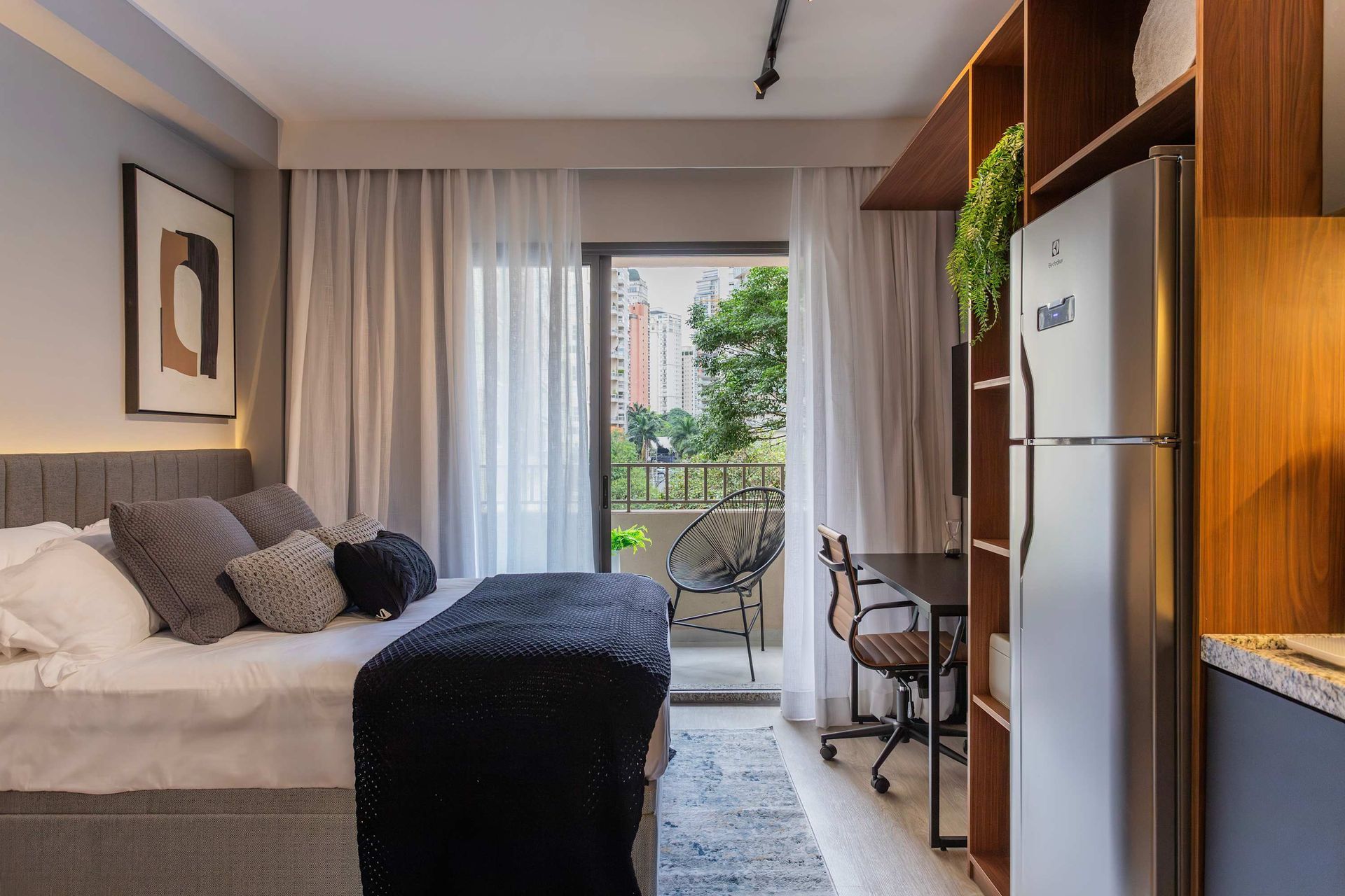 Studio apartment with bed, balcony, desk, refrigerator, and wooden shelving.