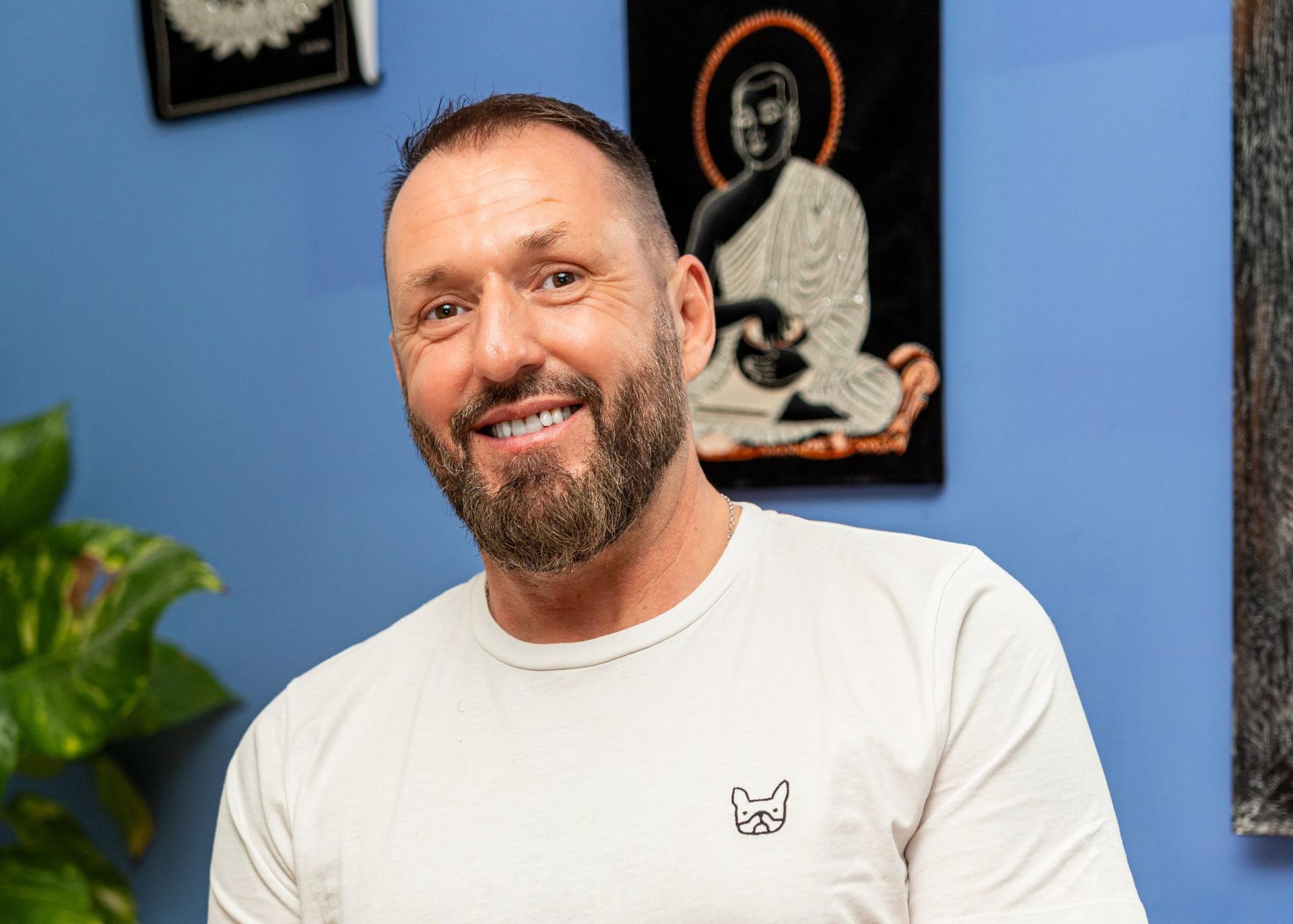 Homme souriant en chemise blanche dans une pièce bleue avec des tableaux encadrés au mur et une plante