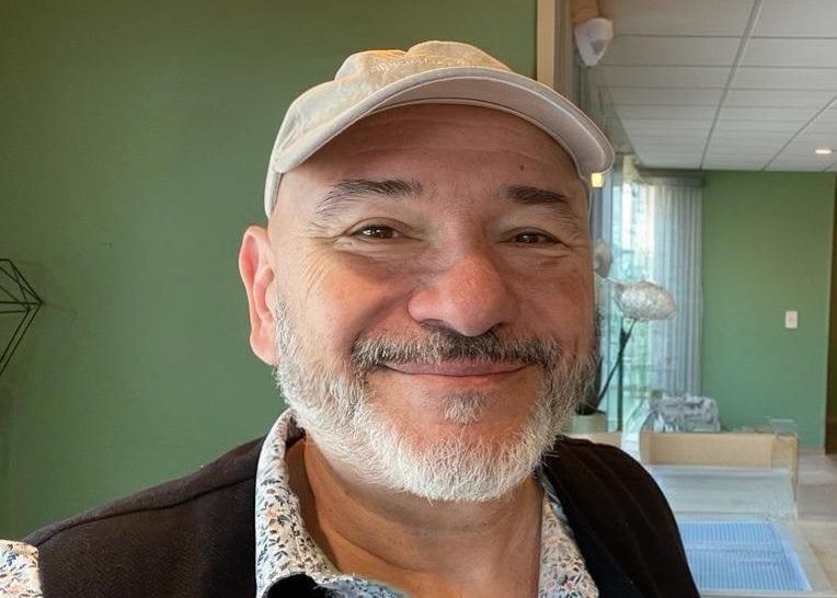 Un homme souriant portant une casquette beige, à l’intérieur, avec un mur vert et une fenêtre en arrière-plan.