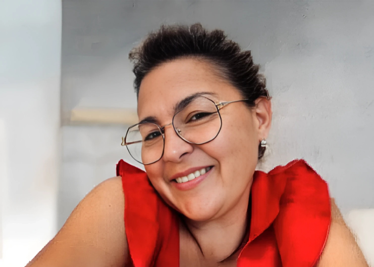 Personne souriante portant des lunettes et un haut rouge, appuyée contre un mur gris clair.