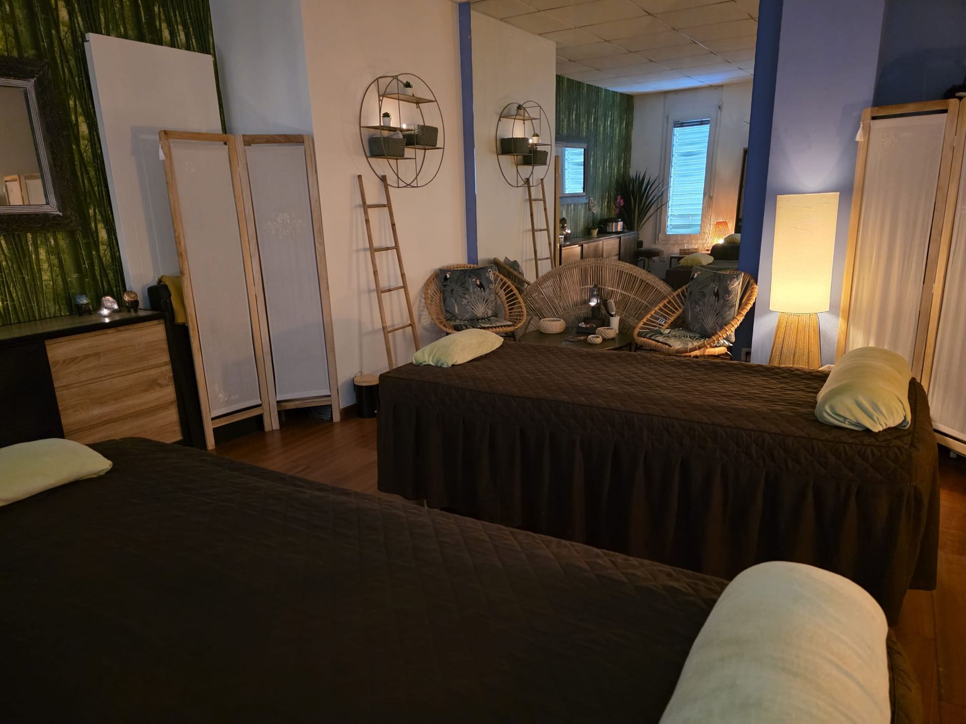 Une chambre douillette avec deux lits, un fauteuil en osier, des miroirs, une échelle et une lumière chaleureuse diffusée par des lampes.