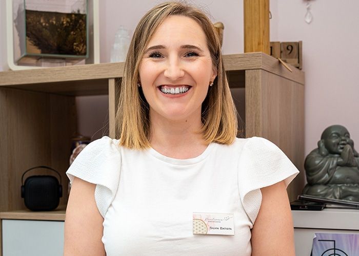 Femme souriante en chemisier blanc debout dans un bureau, avec des étagères et une petite statue derrière elle