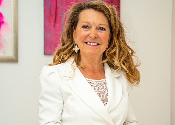 Femme souriante en blazer blanc, debout à l’intérieur à côté d’un tableau abstrait rose.