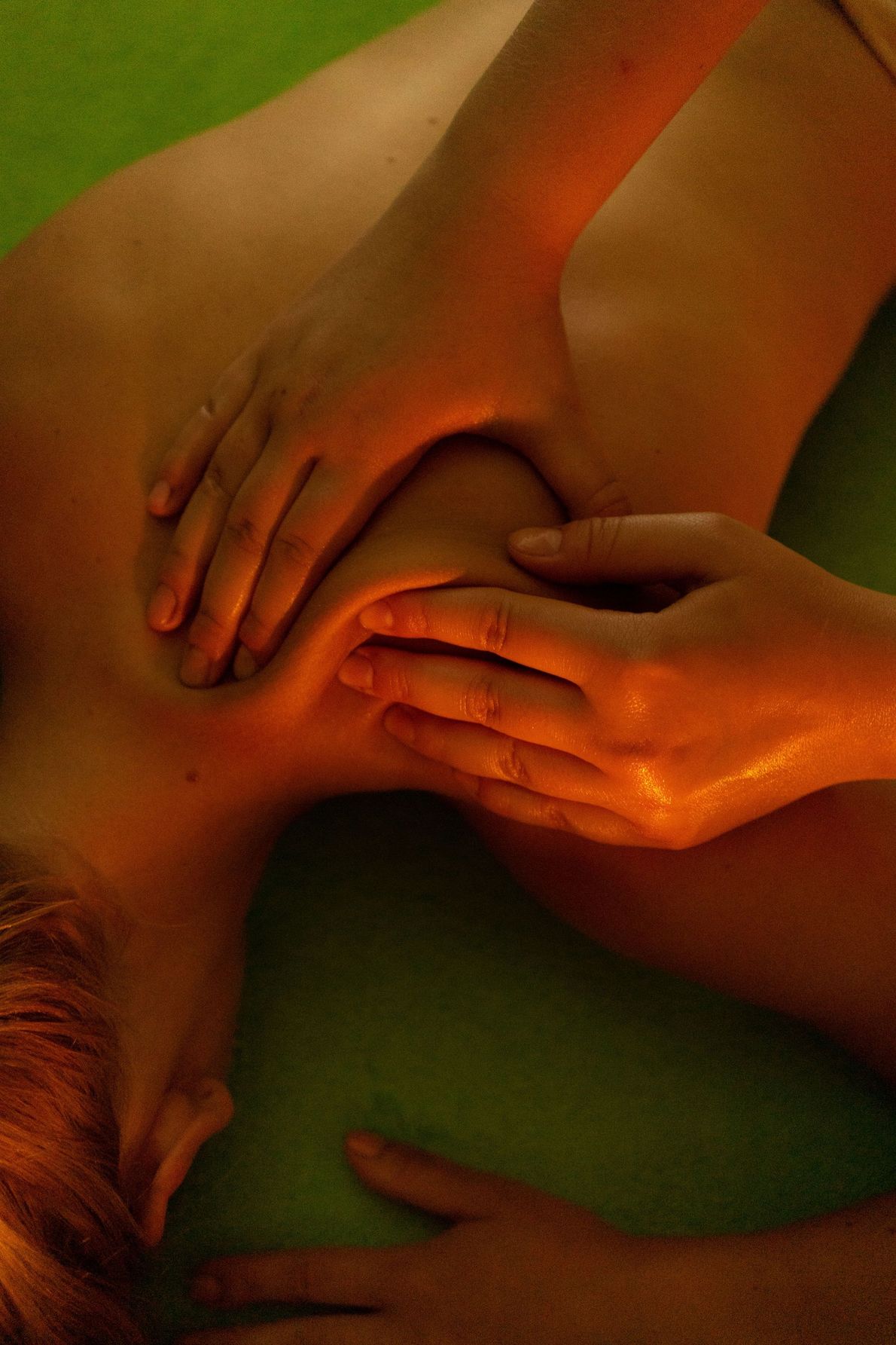 Des mains massagent un dos nu sous une lumière chaude, orange et verte.