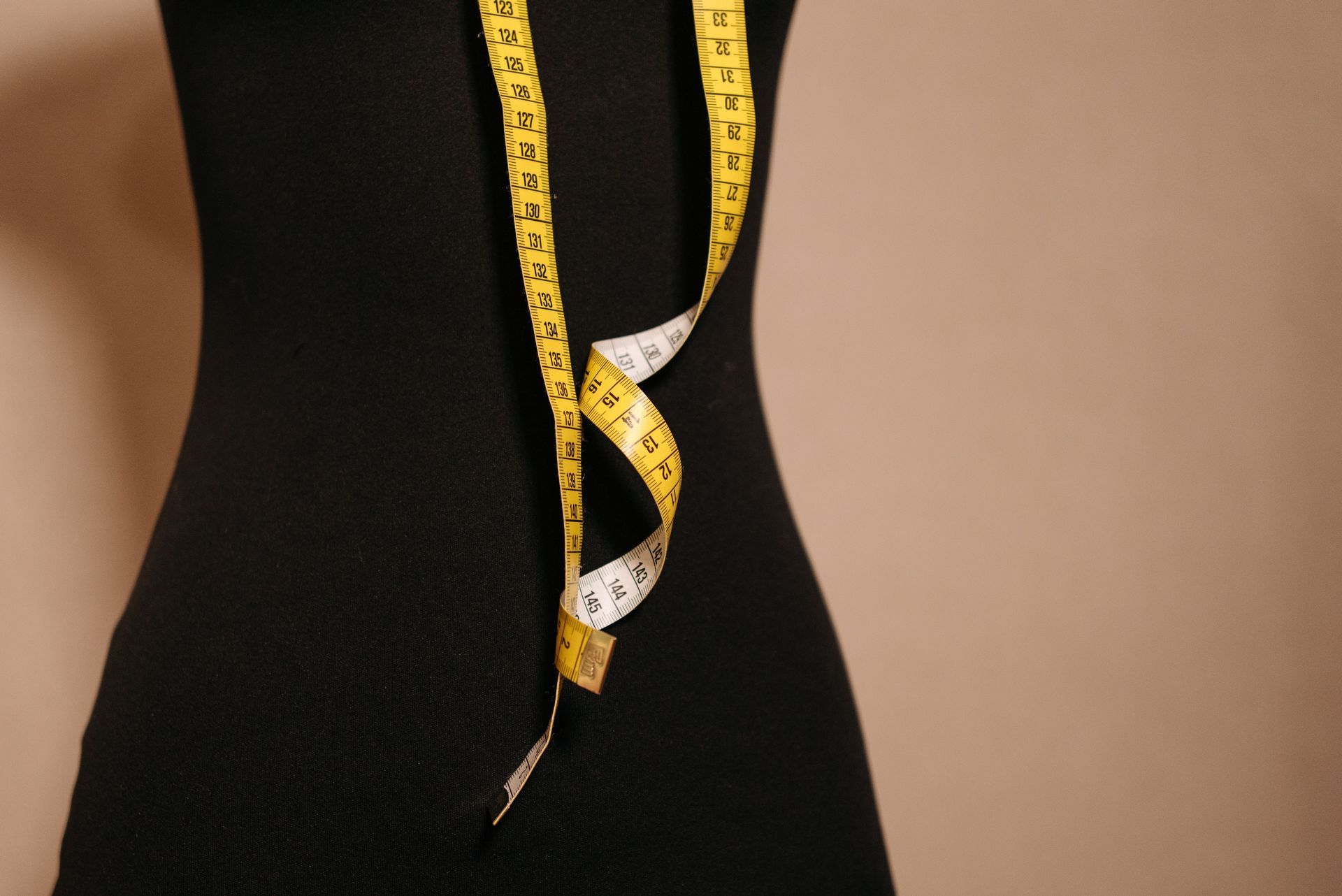 Un mètre ruban jaune enroulé autour d’un mannequin de couture noir sur fond beige