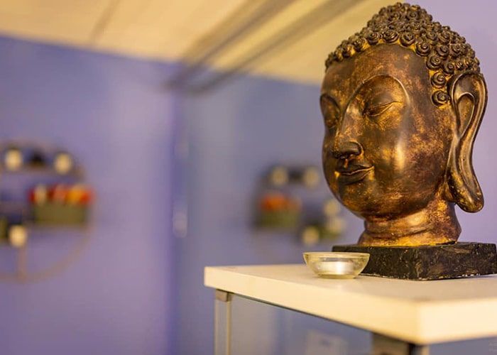 Statue de Bouddha sur une étagère dans une pièce à l’éclairage tamisé, aux murs bleus, avec de petites offrandes à proximité