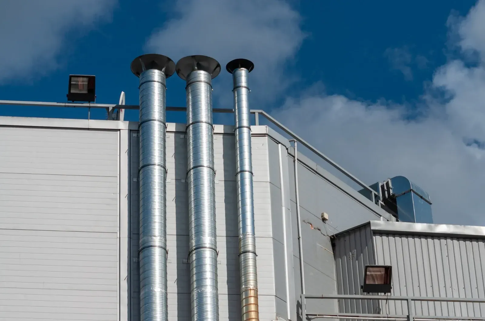 Tres chimeneas metálicas contra un edificio blanco y un cielo azul.
