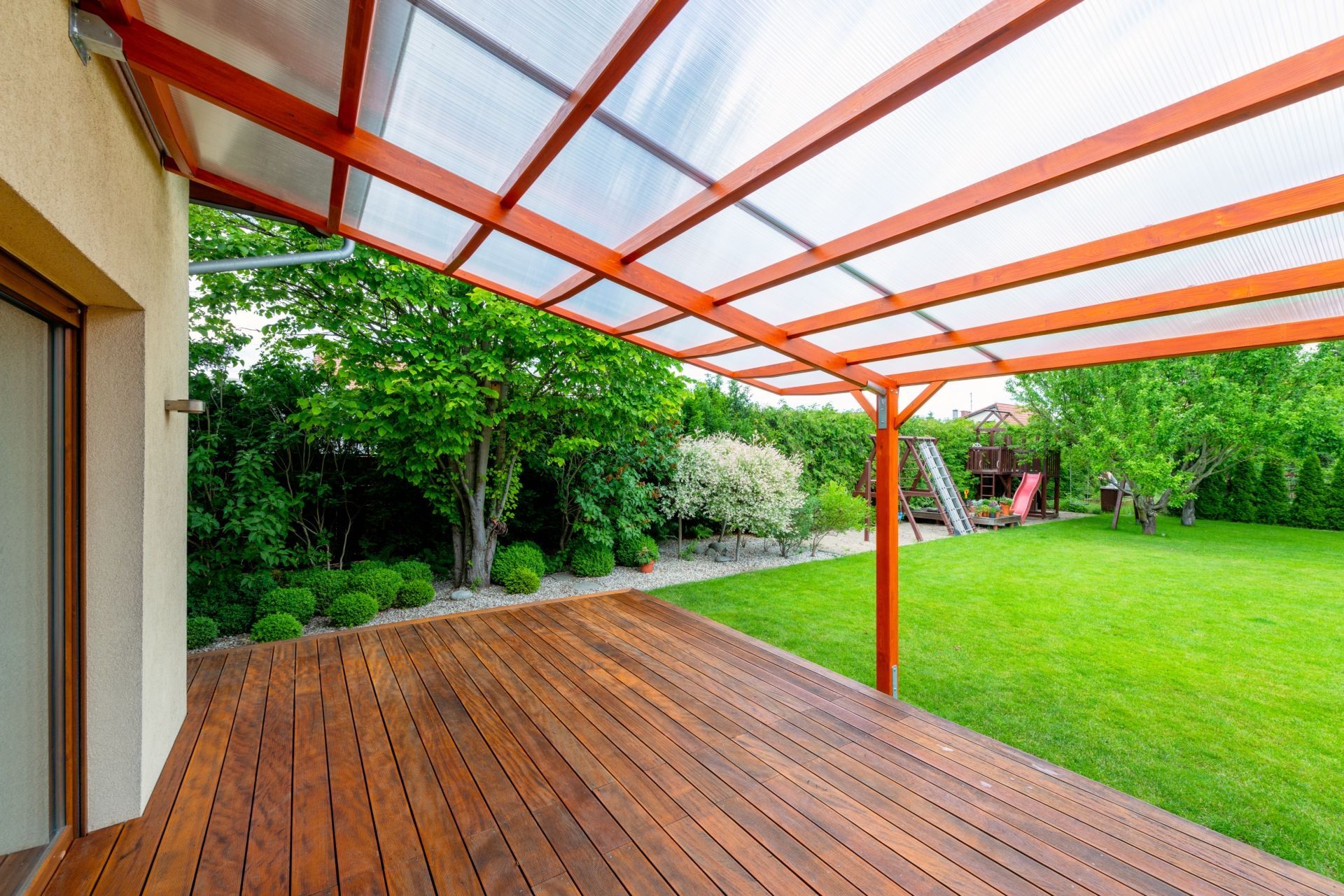 Eine hölzerne Terrasse unter einer rot umrahmten Pergola neben einem grünen Rasen und Garten